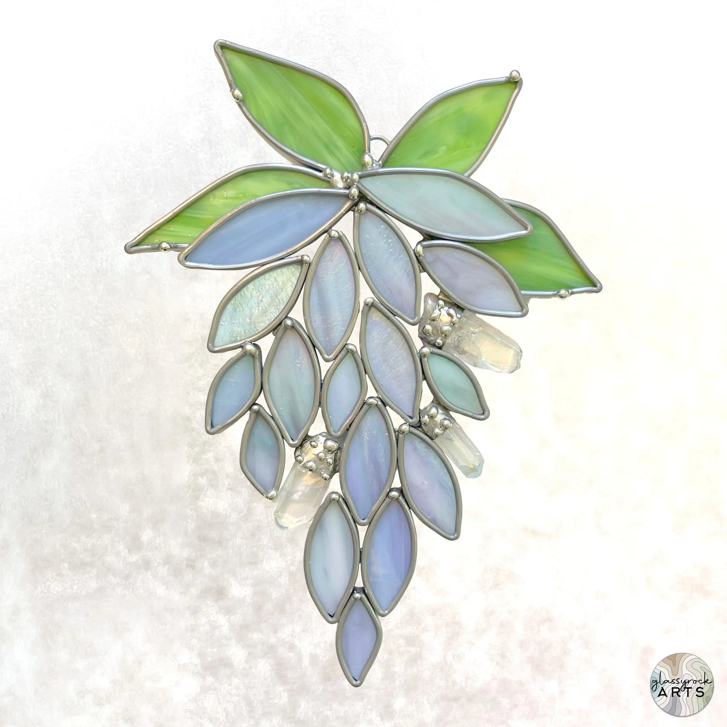 The Handmade Stained Glass Crystal Wisteria Suncatcher features a cluster of leaves and petals in green and blue hues with clear crystal-like accents, artistically arranged against a light background.