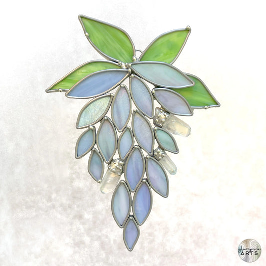 The Handmade Stained Glass Crystal Wisteria Suncatcher features a cluster of leaves and petals in green and blue hues with clear crystal-like accents, artistically arranged against a light background.