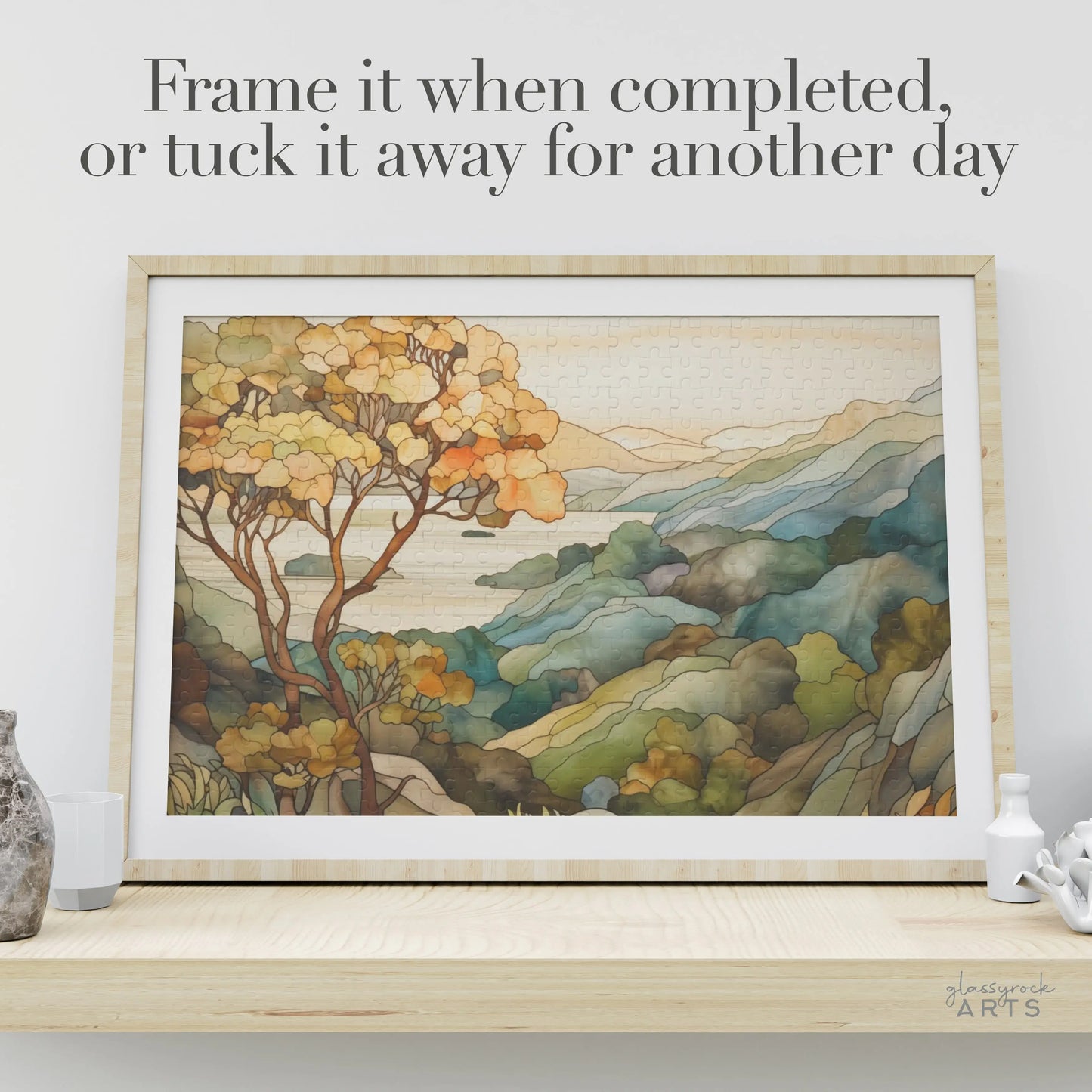 A framed Big Sur California Coast Jigsaw Puzzle of a scenic landscape with trees and hills sits on a wooden shelf. The off-white wall complements the scene, with text above saying, Frame it when completed, or tuck it away for another day.