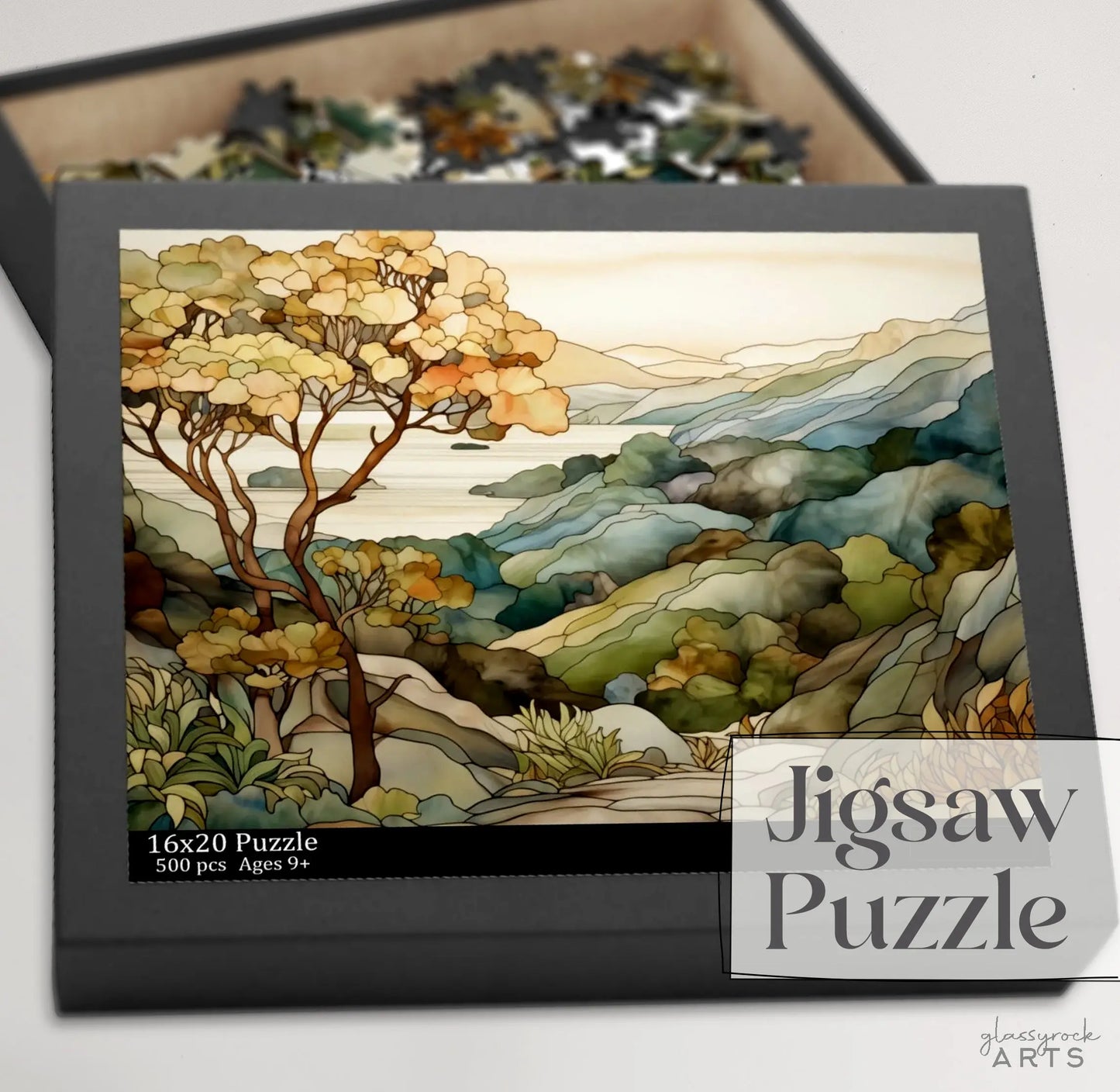 A box of the Big Sur California Coast Jigsaw Puzzle contains 500 pieces depicting a serene nature scene with trees and rolling hills by a lake. The box, marked 16x20 Puzzle and Ages 9+, is ideal as a California coast gift. Puzzle pieces are scattered in an open section of the box.