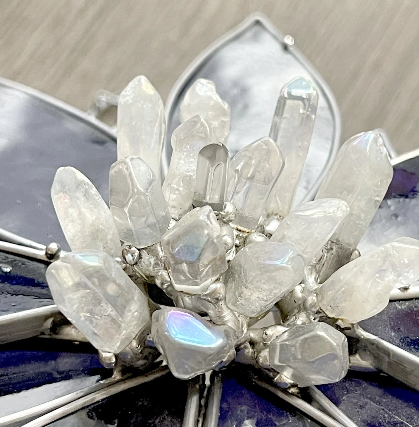 The Handmade Stained Glass Flower and Crystal Suncatcher - XL Blue features a crystal cluster with an iridescent sheen in a metallic frame, resembling flower petals against a bluish-purple backdrop. It beautifully captures light like stained glass, creating an ethereal effect.