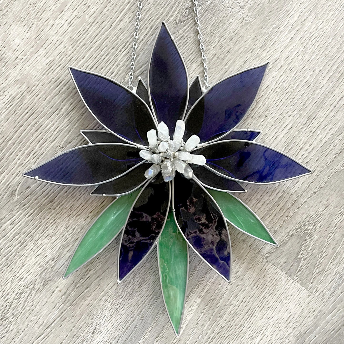 The Handmade Stained Glass Flower and Crystal Suncatcher - XL Blue features dark blue and green flower-shaped petals with a central crystal cluster, hanging from a silver chain on a wooden surface.