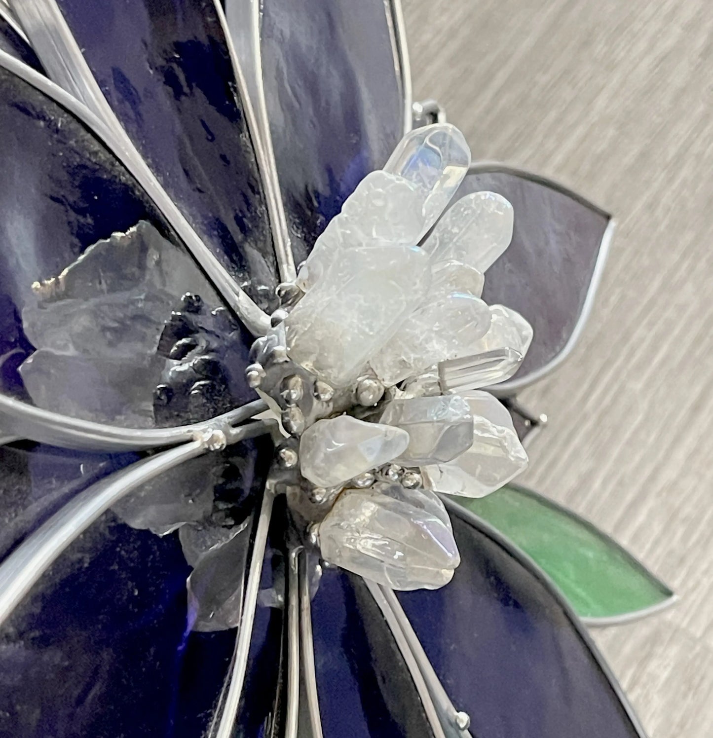 The Handmade Stained Glass Flower and Crystal Suncatcher - XL Blue features a close-up of stained glass flower petals in dark blue and green, centered with a translucent quartz crystal cluster. Its intricate design sits elegantly on a wood-textured surface.
