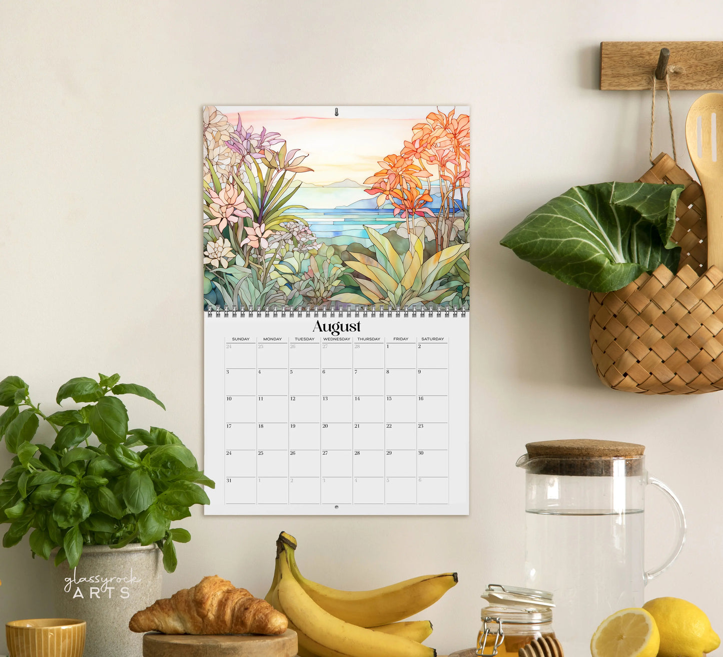A kitchen scene displays a 2025 Botanicals Wall Calendar open to August, featuring a colorful botanical design. Below it sit a potted basil plant, croissants, bananas, a woven basket with utensils, and a glass pitcher. The vibrant calendar enhances the cozy ambiance.