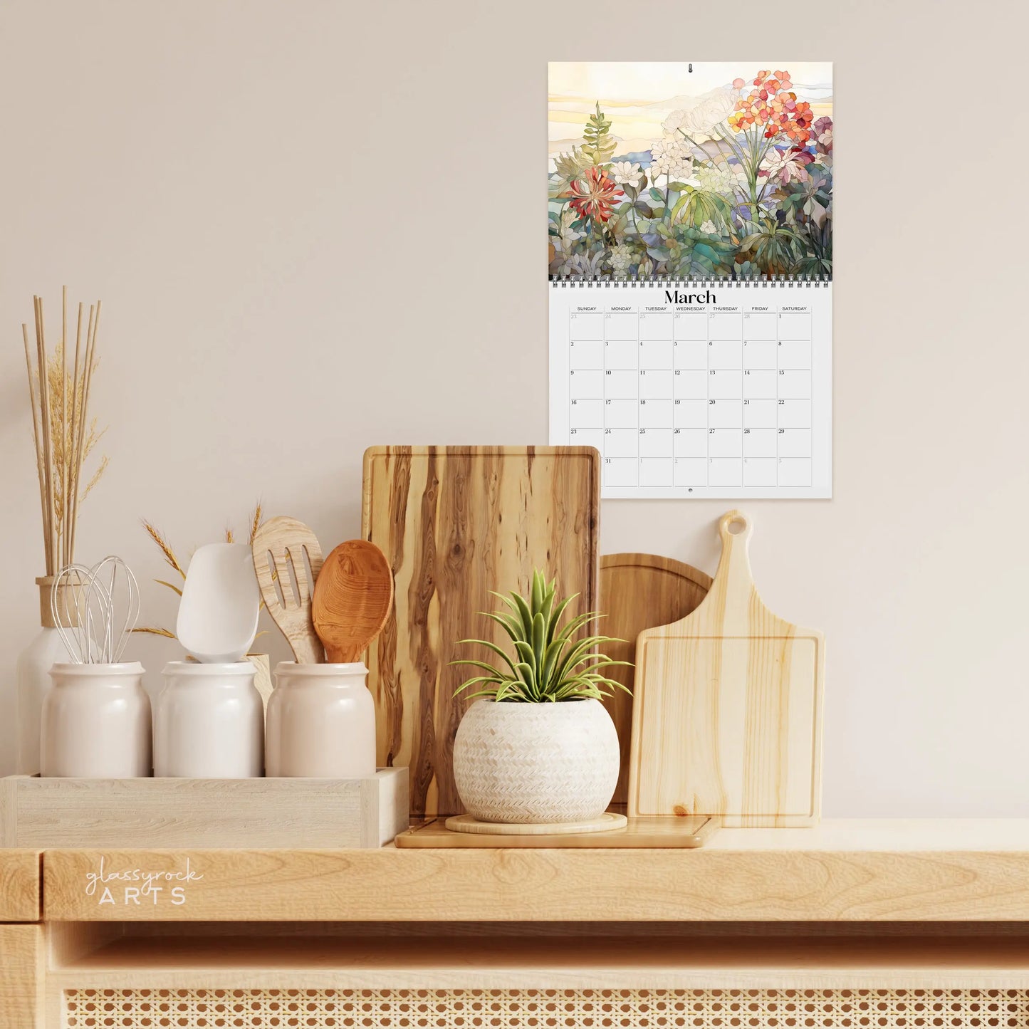 A kitchen counter features a potted plant, wooden utensils, and cutting boards. Above, the 2025 Botanicals Wall Calendar hangs with botanical illustrations on a light-colored background with subtle floral decor.