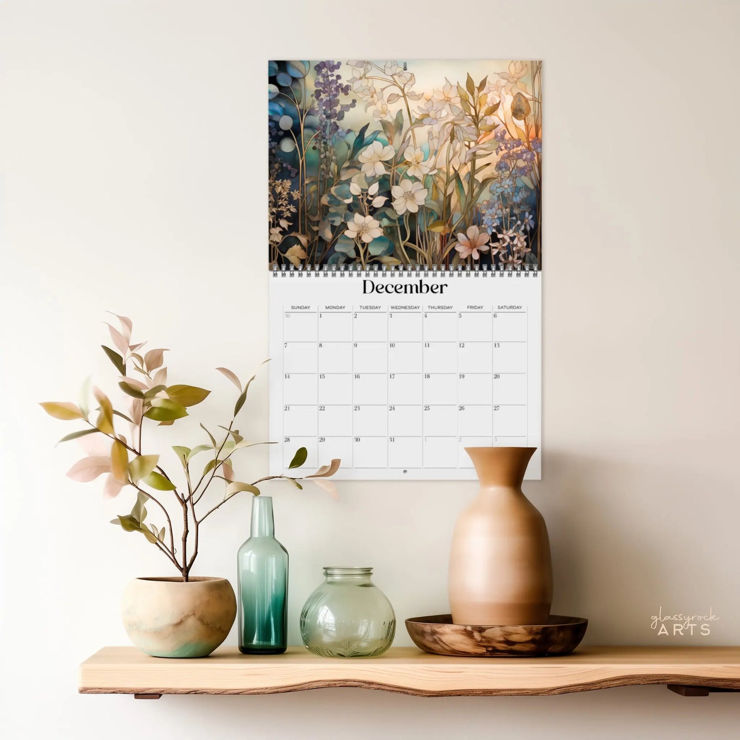 The 2025 Botanicals Wall Calendar for December features stunning botanical illustrations of white and blue flowers, hanging above a shelf adorned with decorative vases and a small plant against a light-colored wall.