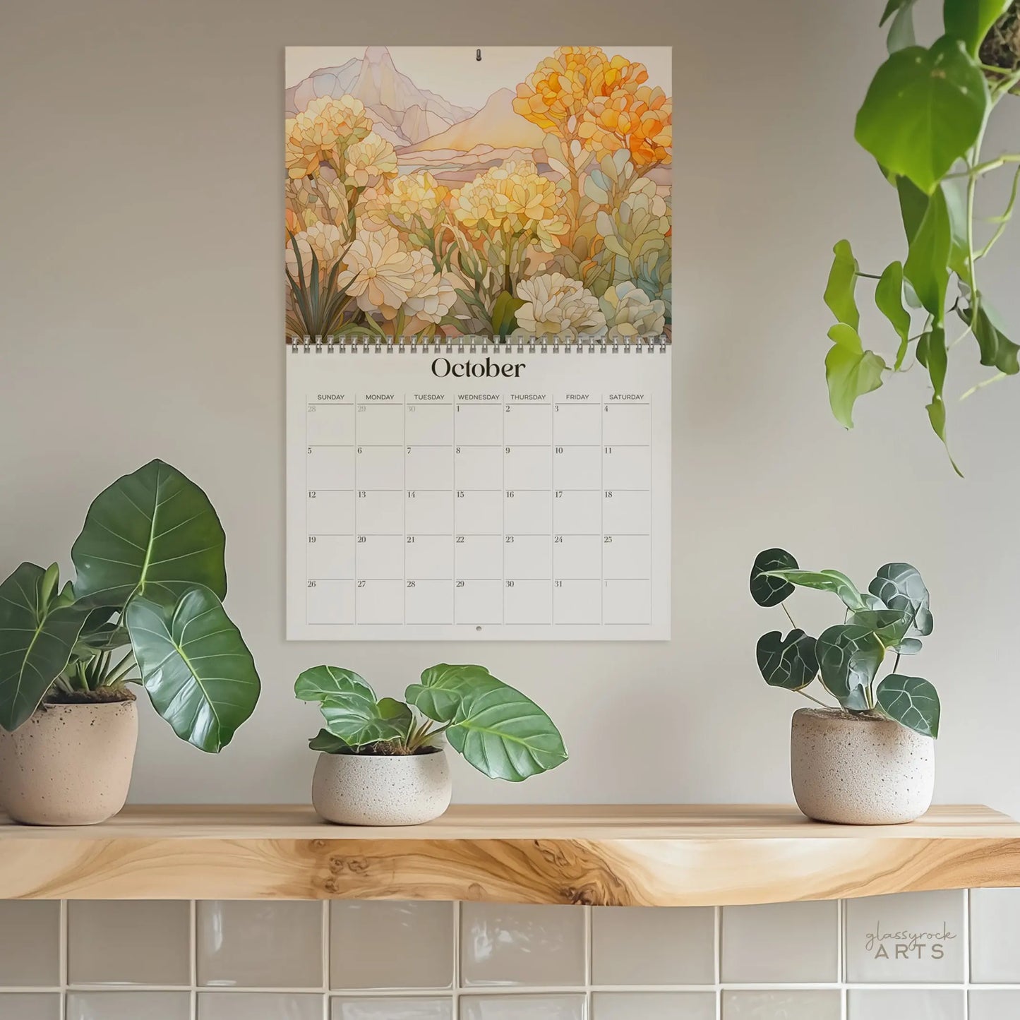 The 2025 Botanicals Wall Calendar is open to October, showing mountains with orange and white flowers. Below it, three potted plants rest on a light wooden shelf, along with a hanging plant on the right.