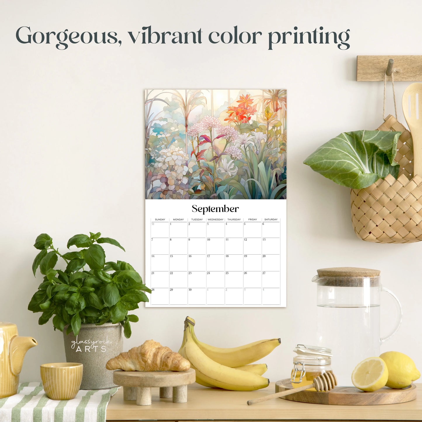 A serene kitchen scene showcases the 2025 Botanicals Wall Calendar, displaying stunning botanical illustrations. On the counter are a plant, woven basket with cabbage, wooden cutting board, glass pitcher, and bananas. Text reads, Gorgeous, vibrant color printing.