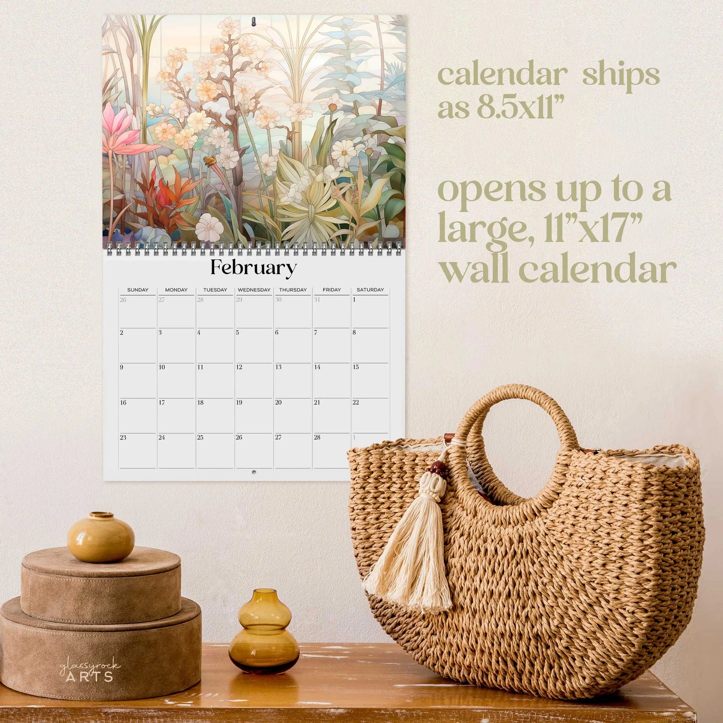 A decorative wall display features the 2025 Botanicals Wall Calendar open to February, next to a tasseled woven basket and stacked round containers. The calendar ships as 8.5x11 and expands to an impressive 11x17.