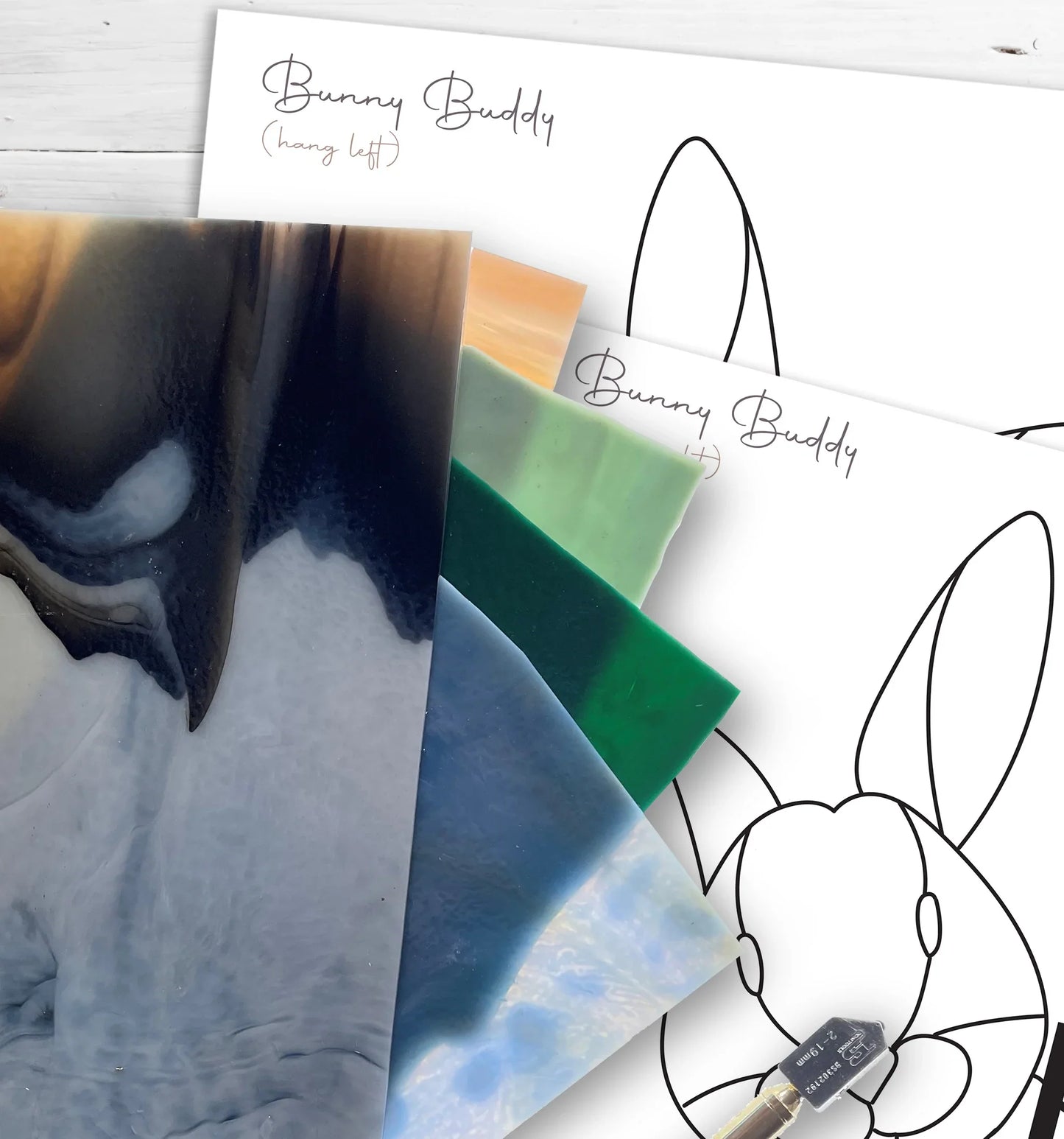 The Animal Stained Glass Pattern 9-Pack includes glass sheets in dark blue, green, and white atop Bunny Buddy outline drawings. Ideal for intermediate crafters, a small brush awaits your next creative touch.