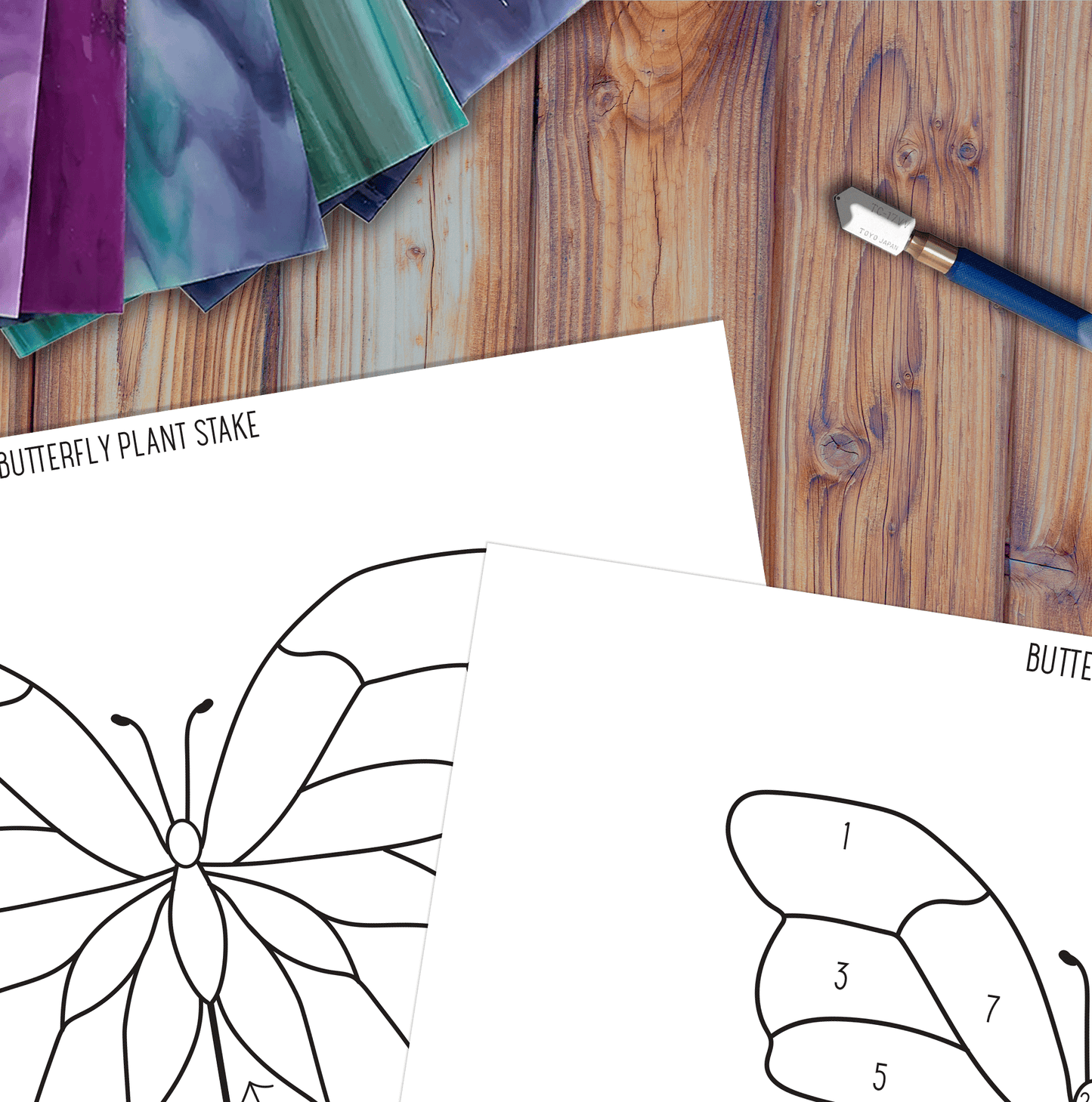 Colorful stained glass sheets are set on a wooden surface with butterfly crafting templates and a knife, ideal for making a stunning butterfly garden stake using the Butterfly Garden Stake Stained Glass Pattern digital download.