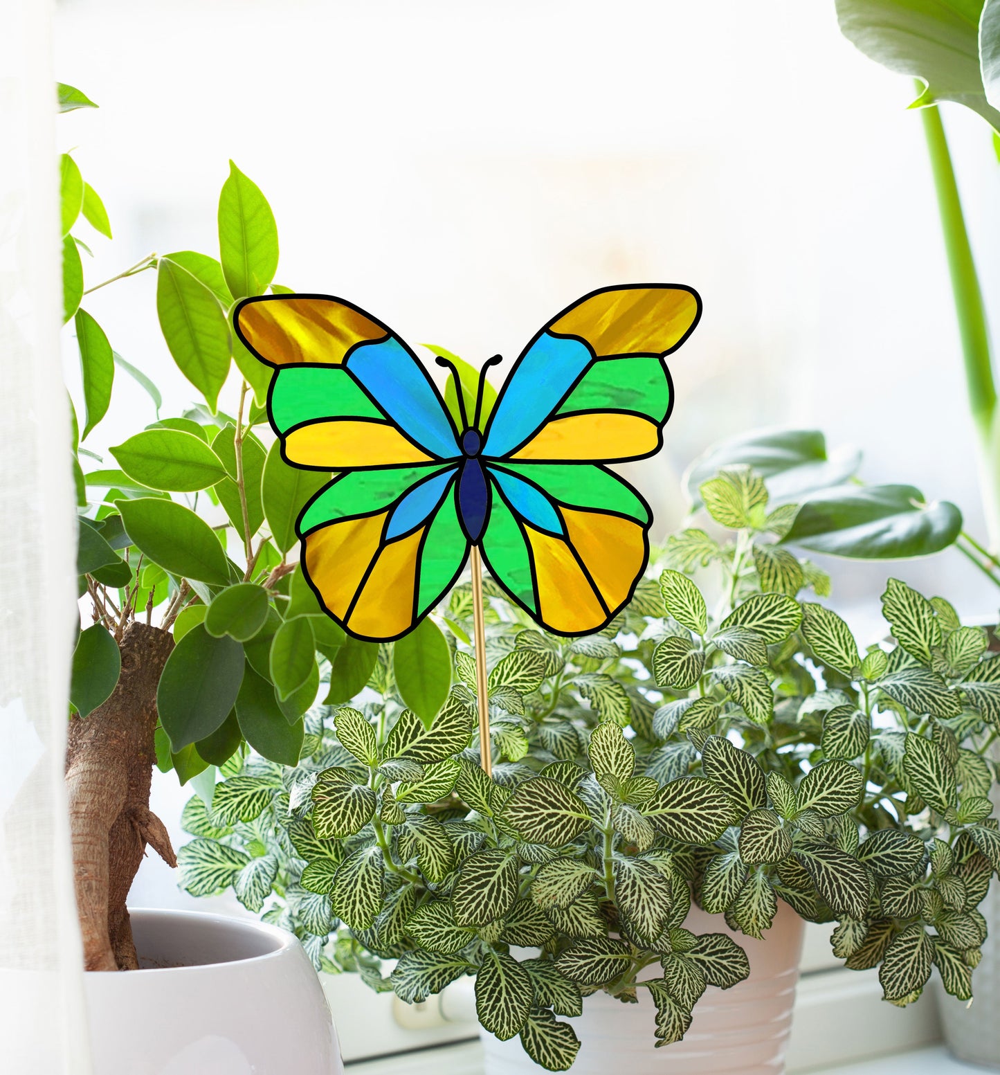 The Butterfly Garden Stake Stained Glass Pattern sits among the lush green leaves of potted plants on a windowsill, with sunlight filtering through the window to illuminate its intricate blue, yellow, and green hues.