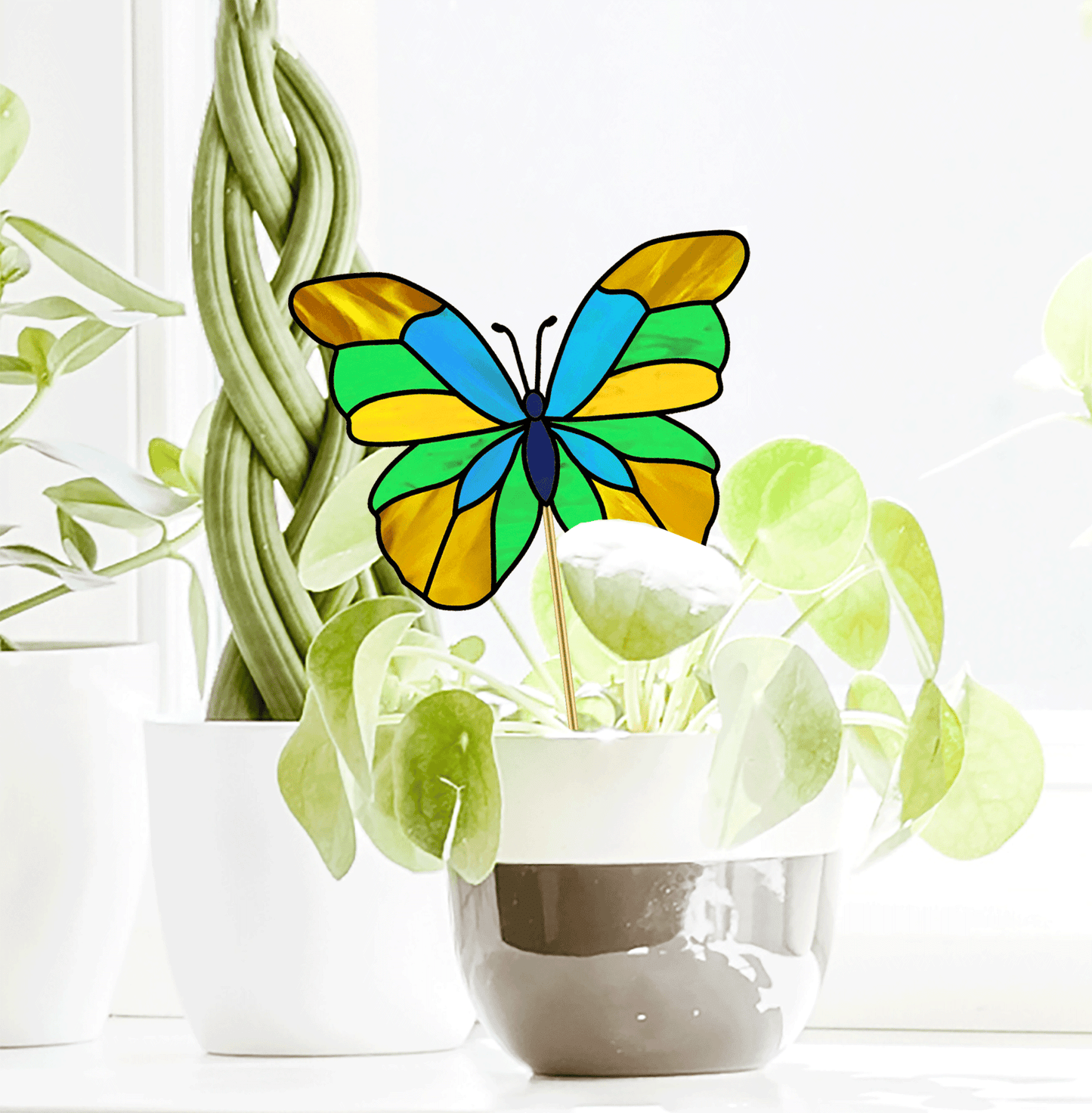 A stained glass butterfly with vibrant green, yellow, and blue wings perches among plants on a sunlit windowsill. Enhance your decor with this beauty or explore our Butterfly Garden Stake Stained Glass Pattern download to craft your own masterpiece.