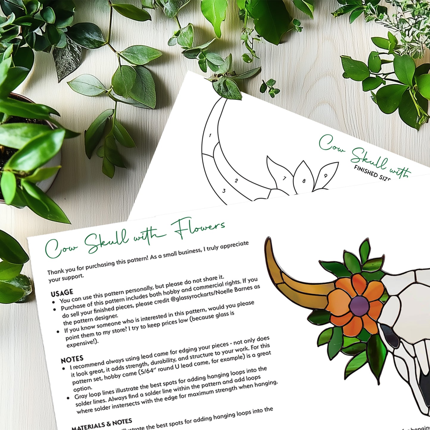 The Cow Skull Floral Stained Glass Pattern lies on a wooden surface, resembling a cow skull suncatcher. It features instructions, a partially colored floral skull with scattered leaves, and is adorned with green plants to enhance its natural theme.