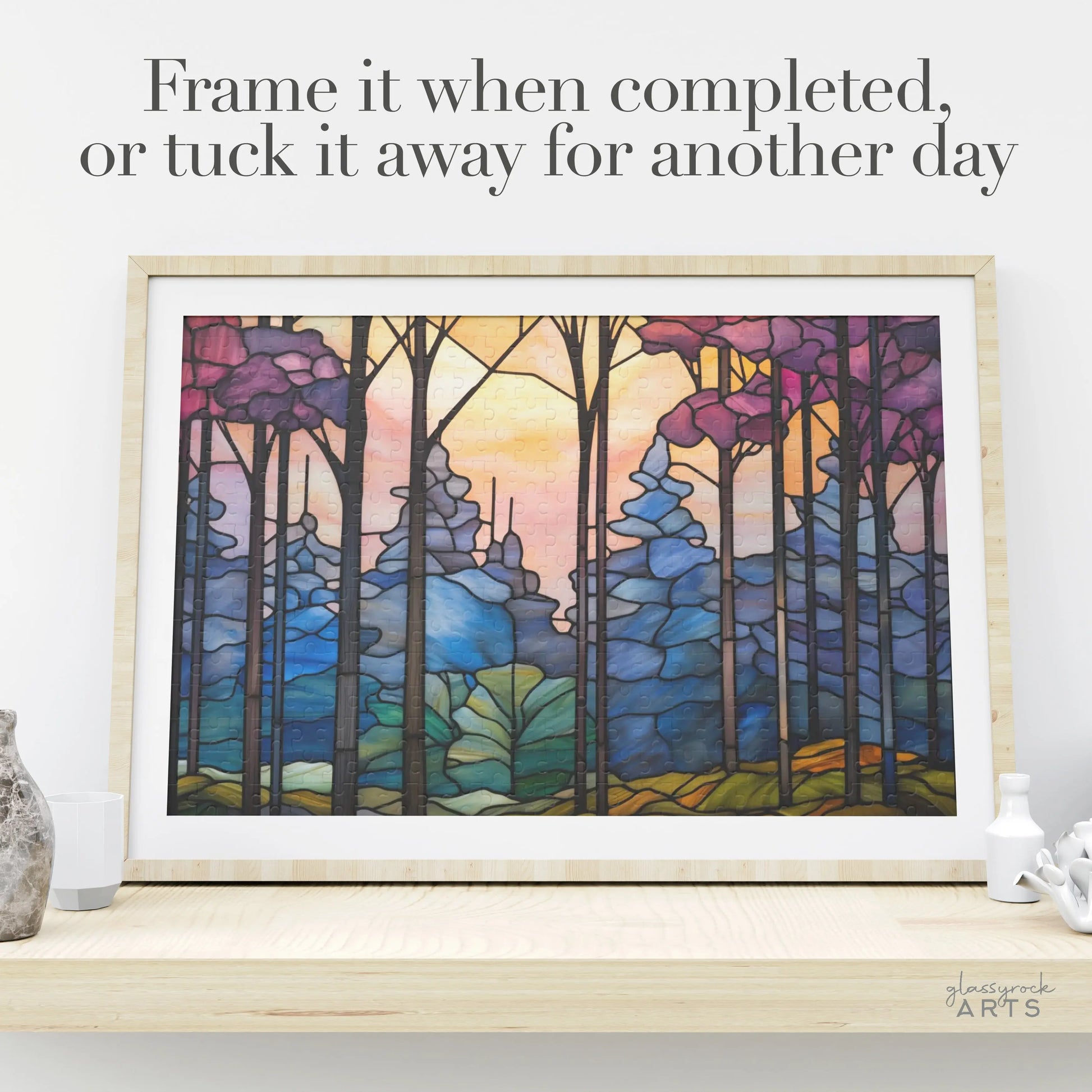 The Dark Woods Forest Jigsaw Puzzle, depicting a colorful forest with abstract trees and a sunset sky, sits on a light-colored shelf beside decorative vases. Text above suggests, Frame it when completed, or tuck it away for another day. Its the perfect art gift for any occasion.