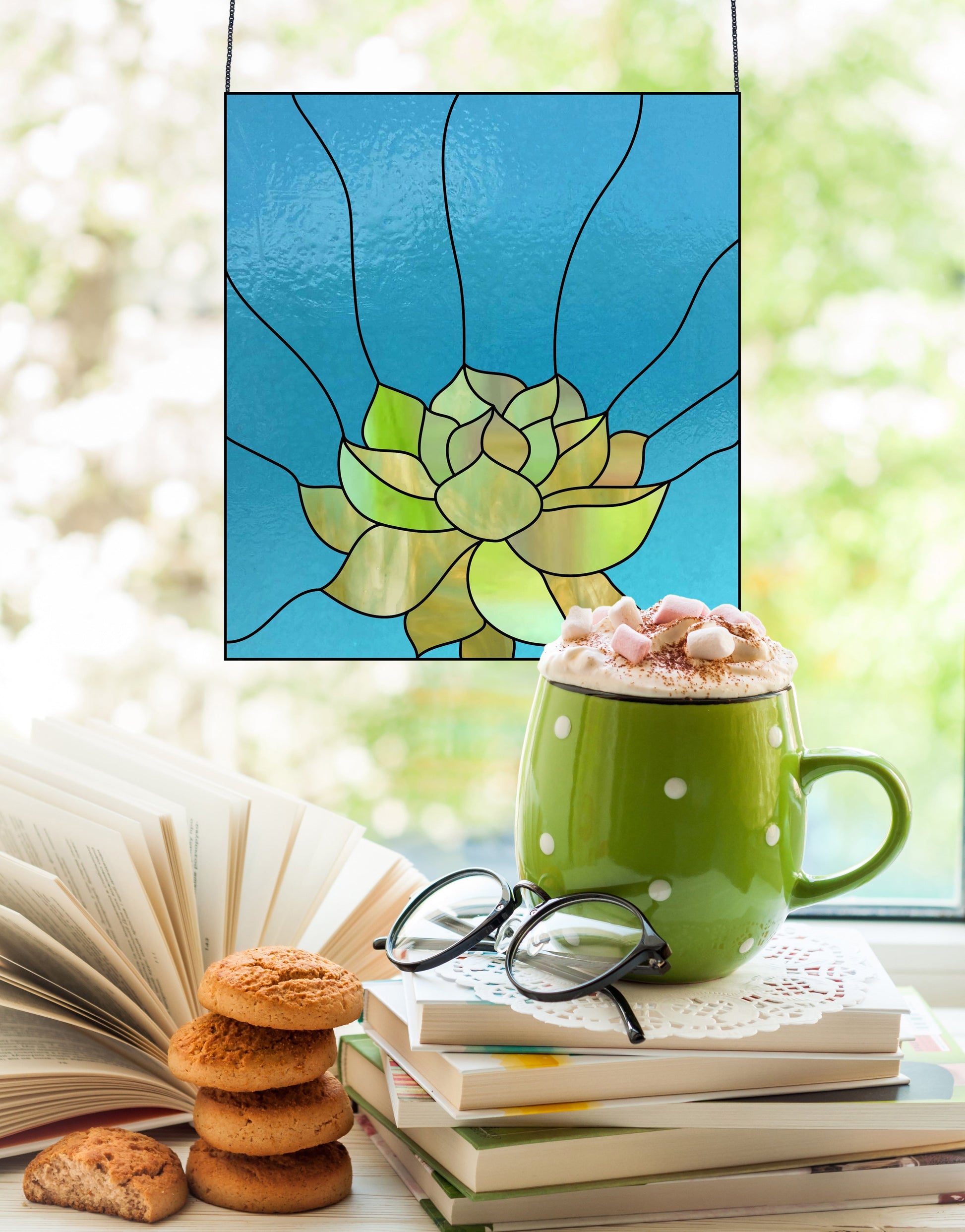 A green mug of hot chocolate with marshmallows sits on books and glasses beside cookies. Through the window, a floral Stained Glass Succulent Panel Pattern casts vibrant patterns, revealing glimpses of the blurred garden beyond.