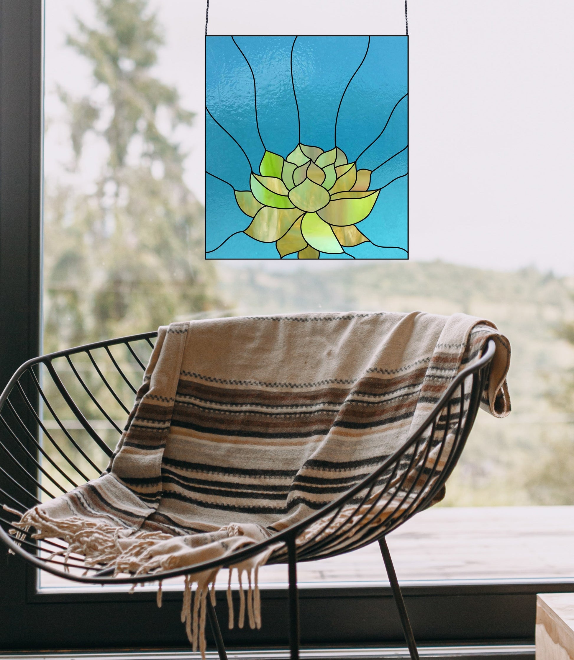 In front of a window, a modern black wire chair with a striped blanket stands, adorned by the Stained Glass Succulent Panel Pattern. This artwork features green and yellow hues on blue glass, offering a serene view of trees and hills stretching beyond it.