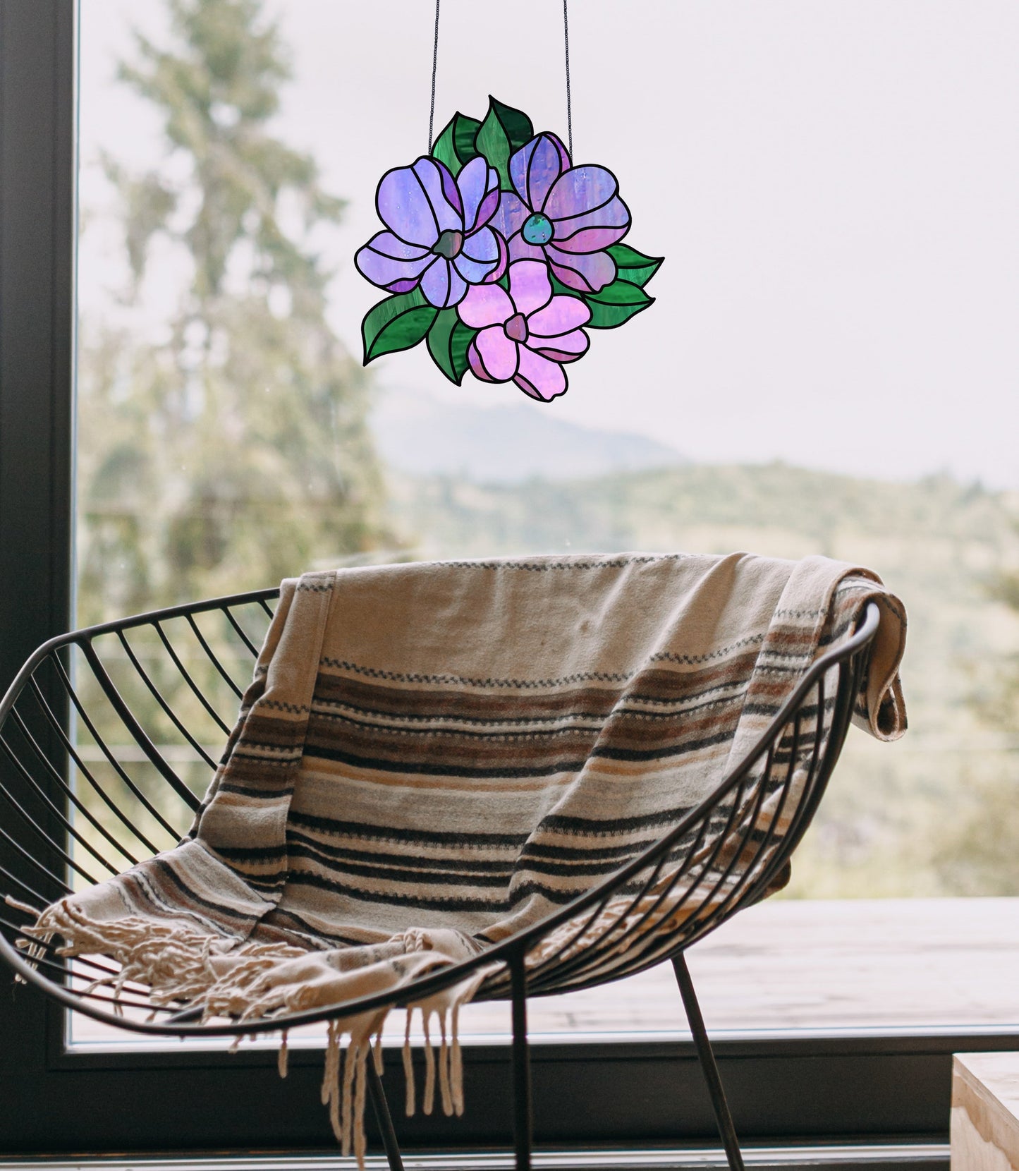 A black wire chair with a striped throw blanket sits near the window. A stunning Bundle of Flowers Stained Glass Pattern featuring purple and pink blooms with green leaves hangs in the foreground, offering a scenic view of trees and hills outside.