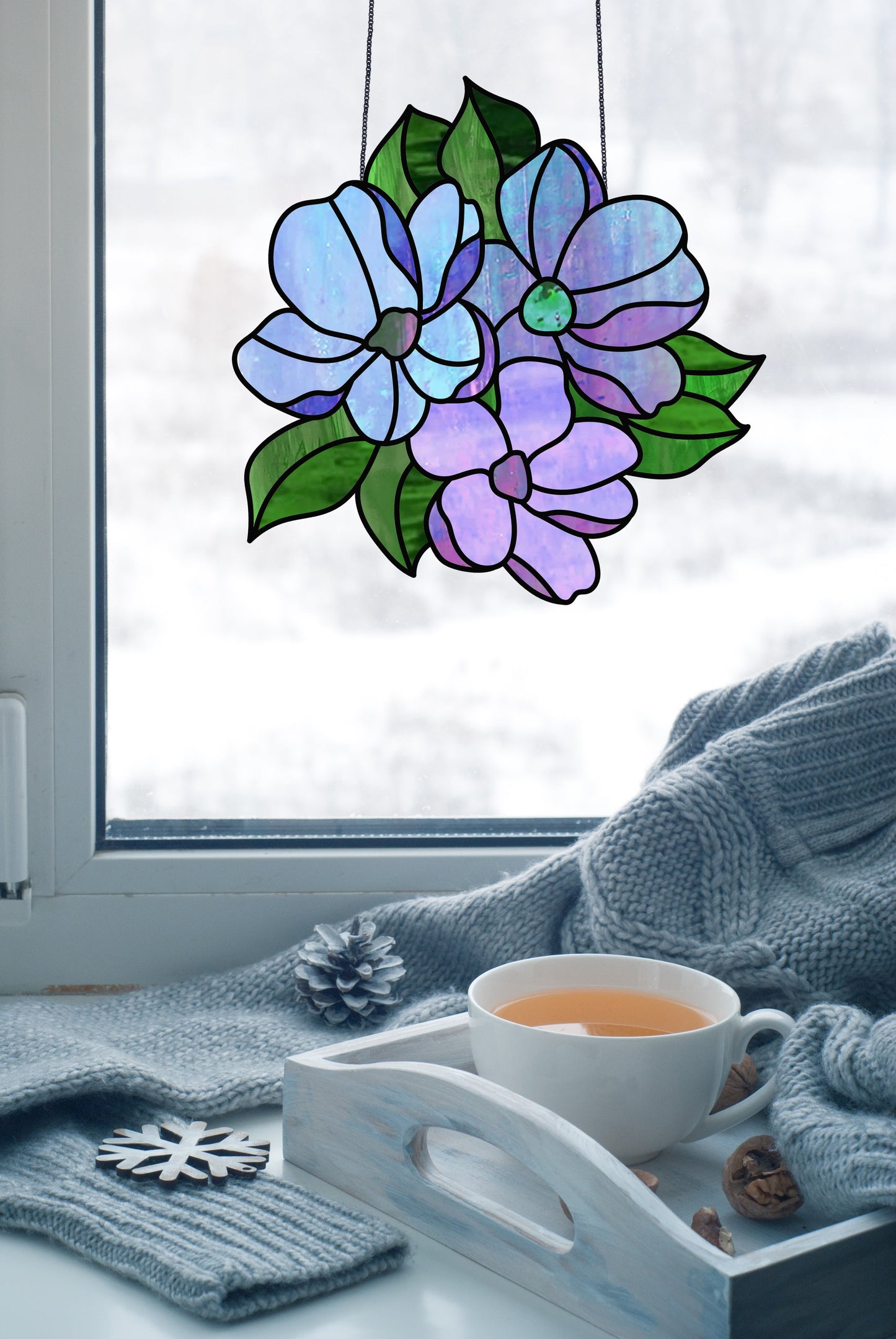 The Bundle of Flowers Stained Glass Pattern suncatcher hangs by the window, casting colors against a snowy scene. In front, a tray displays a cup of tea, cinnamon sticks, pine cone, and wooden snowflake on a cozy gray blanket.