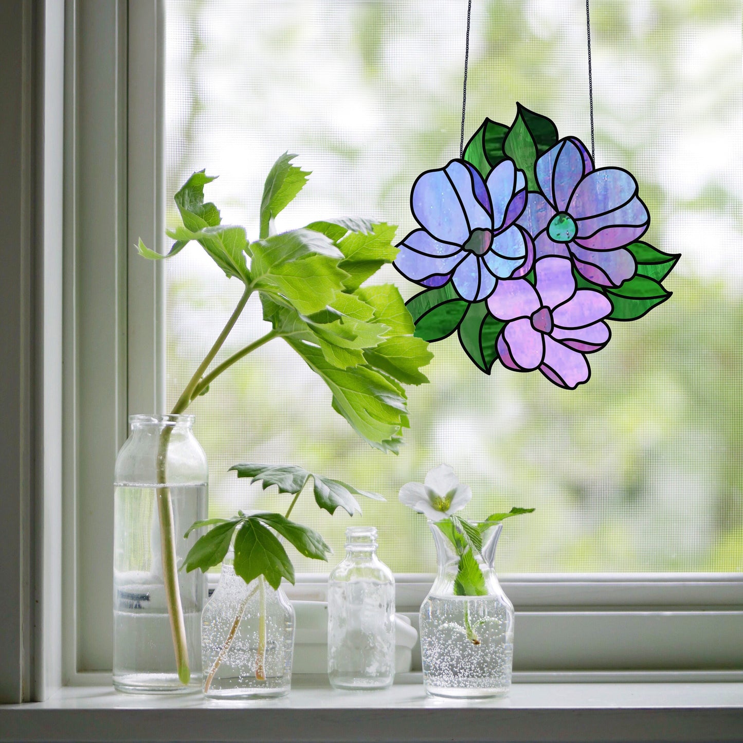 Three glass bottles with green leaves and a white flower stand on a windowsill. Nearby, a Bundle of Flowers Stained Glass Pattern suncatcher casts vibrant reflections. The background is a soft, natural view through the blurred window.