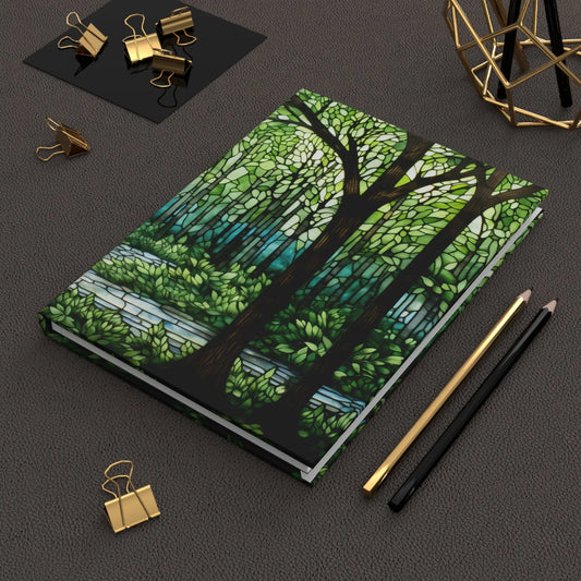 The Stained Glass Forest Hardcover Journal showcases a unique stained glass design on a textured gray cover. Complete your creative setup with gold and black pencils, gold binder clips, and a geometric decorative object—ideal companions for your artistic explorations.