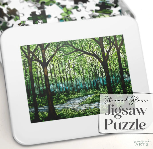 The Stained Glass Forest Jigsaw Puzzle box features a forest scene with tall trees and a river in stained glass style, surrounded by scattered pieces. It makes an exquisite gift for any nature lover.