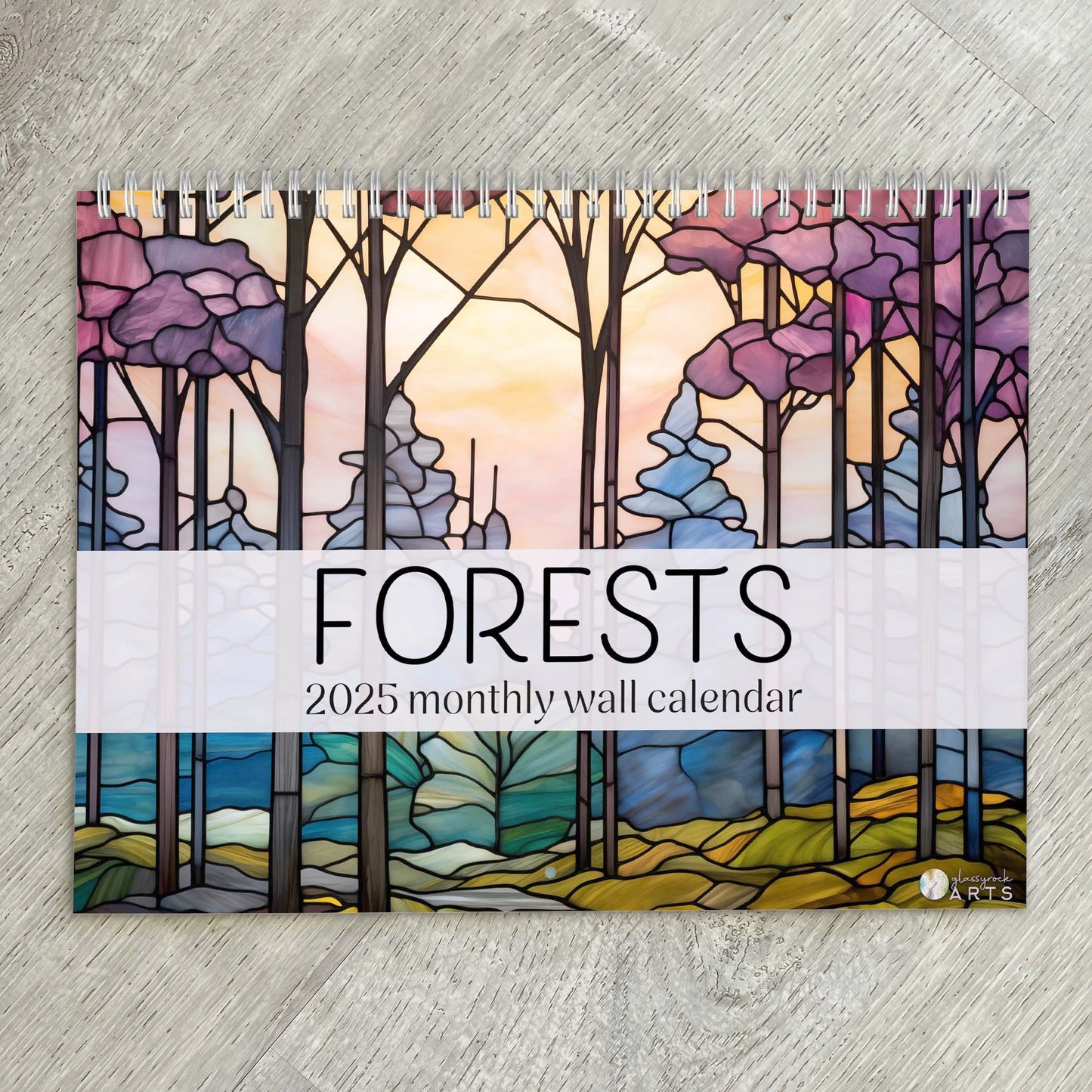 The image features the 2025 Forests Stained Glass Wall Calendar with vibrant tree illustrations. Spiral-bound, it rests elegantly on a wooden surface.