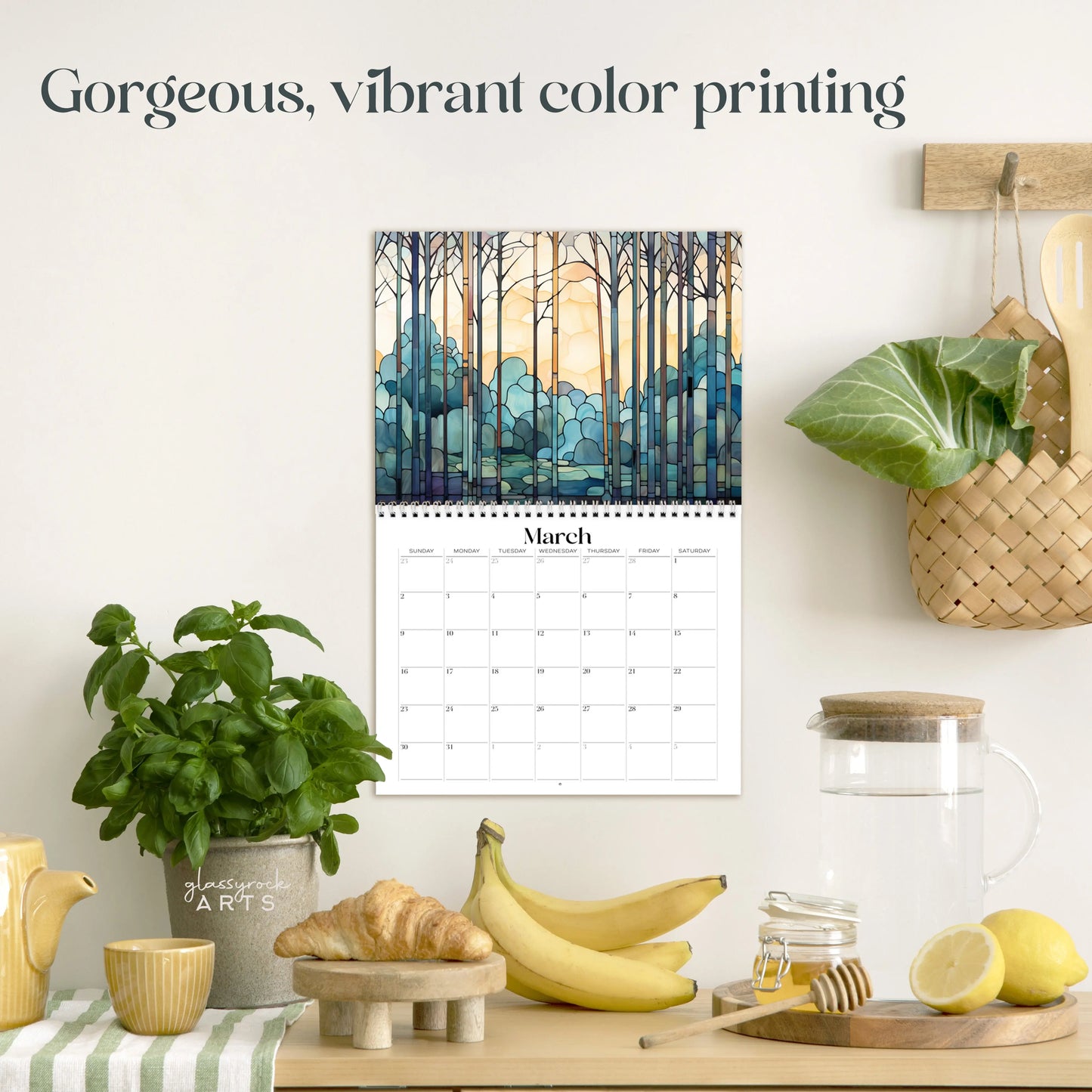In a kitchen scene, the 2025 Forests Stained Glass Wall Calendar hangs elegantly on the wall. Below it, potted herbs, bananas, a glass pitcher, and a woven basket adorn the light countertop. The text proudly states: Gorgeous, vibrant color printing.