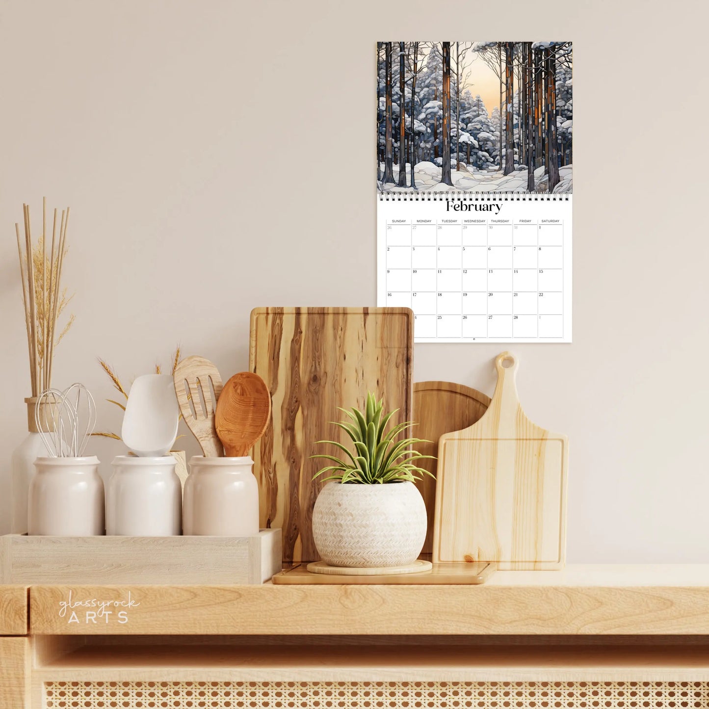 A kitchen counter showcases a potted succulent, cooking utensils, and wooden cutting boards. Above, the 2025 Forests Stained Glass Wall Calendar depicts a snowy forest with trees and a sunset sky, creating a cozy and natural ambiance highlighted by the calendars charming nature art.