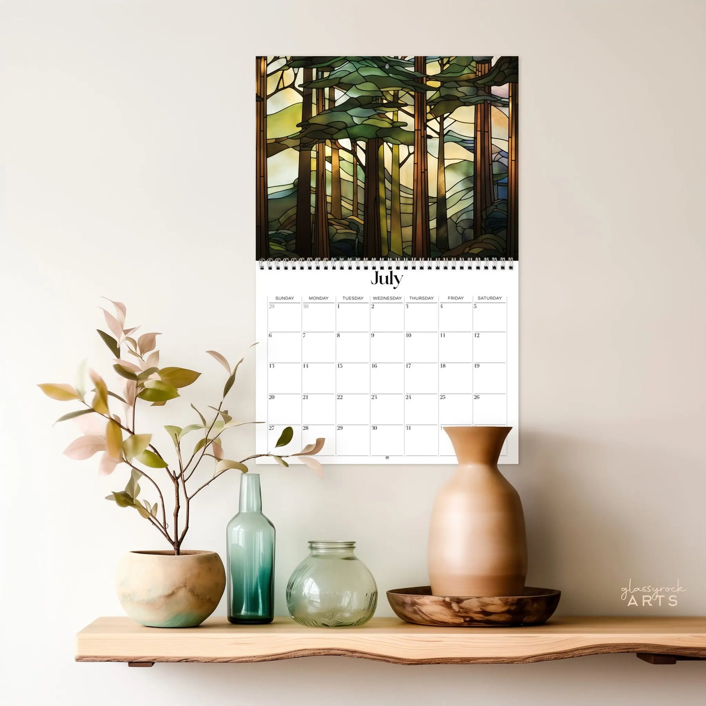 A 2025 Forests Stained Glass Wall Calendar showing July hangs above a wooden shelf. It depicts an artistic forest scene with tall trees and stained glass effects. The shelf holds decorative vases and a small plant.