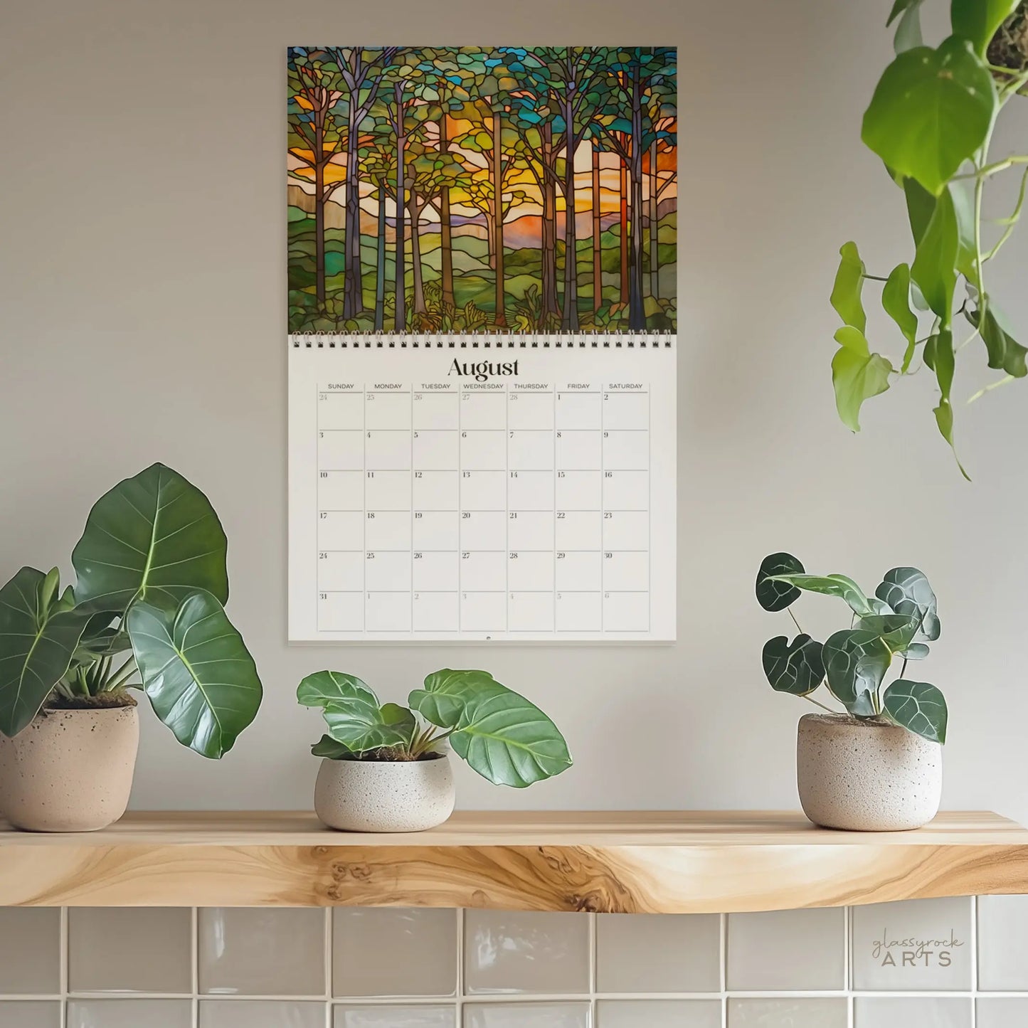 A 2025 Forests Stained Glass Wall Calendar with a vibrant forest theme hangs on a light wall. Below, three potted plants rest on a wooden shelf, while one leafy plant dangles from above, infusing greenery into the setting.