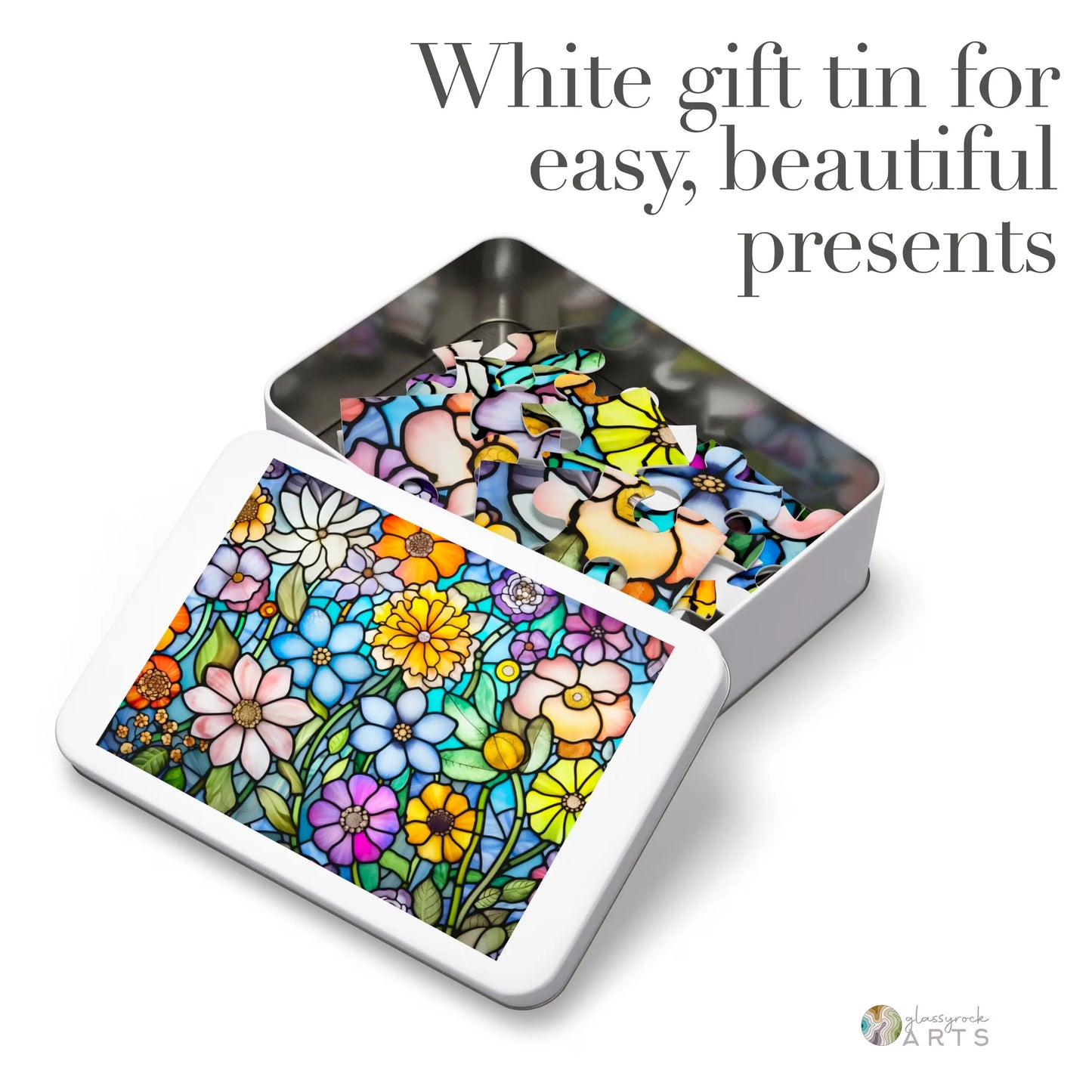 A white gift tin is partially opened, displaying the Stained Glass Flower Garden Jigsaw Puzzle with a vibrant floral pattern. The lid features the same design. Text reads, Stained glass puzzle gift: white tin for elegant presents made from high-quality chipboard.