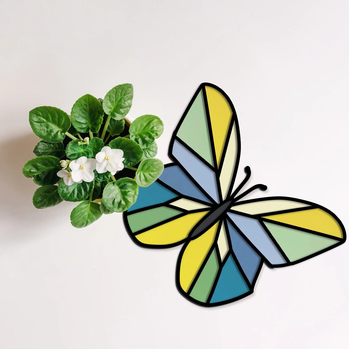 A white-flowered potted plant sits beside a vibrant geometric stained glass butterfly pattern, showcasing yellow, green, and blue shades on a white surface. (Product: Stained Glass Geometric Butterfly SVGs).