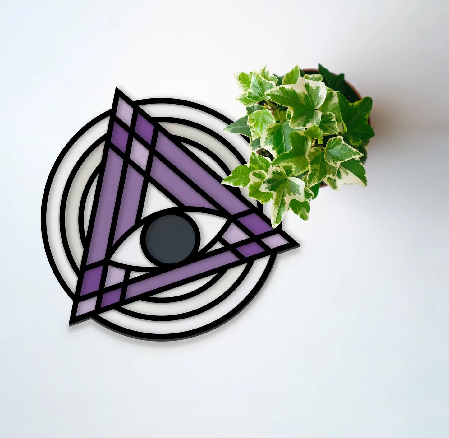 A potted ivy plant rests on a white surface beside a geometric stained glass artwork named Stained Glass Evil Eye Suncatcher SVGs, featuring a purple triangle with a central circular black shape, surrounded by black and white rings.