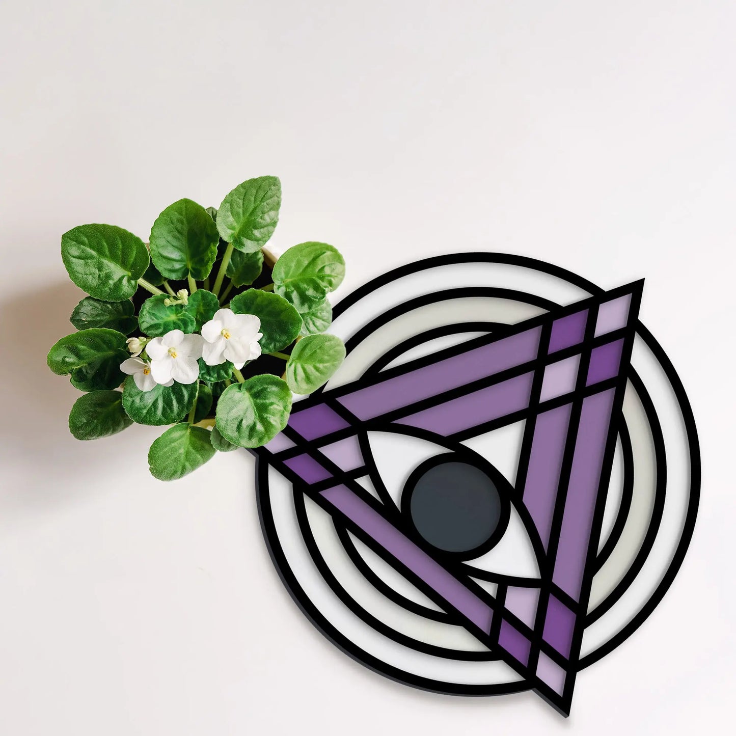 A potted plant with green leaves and white flowers sits on a light surface beside a Stained Glass Evil Eye Suncatcher SVG design, featuring a triangle and concentric circles in black, lavender, and white.