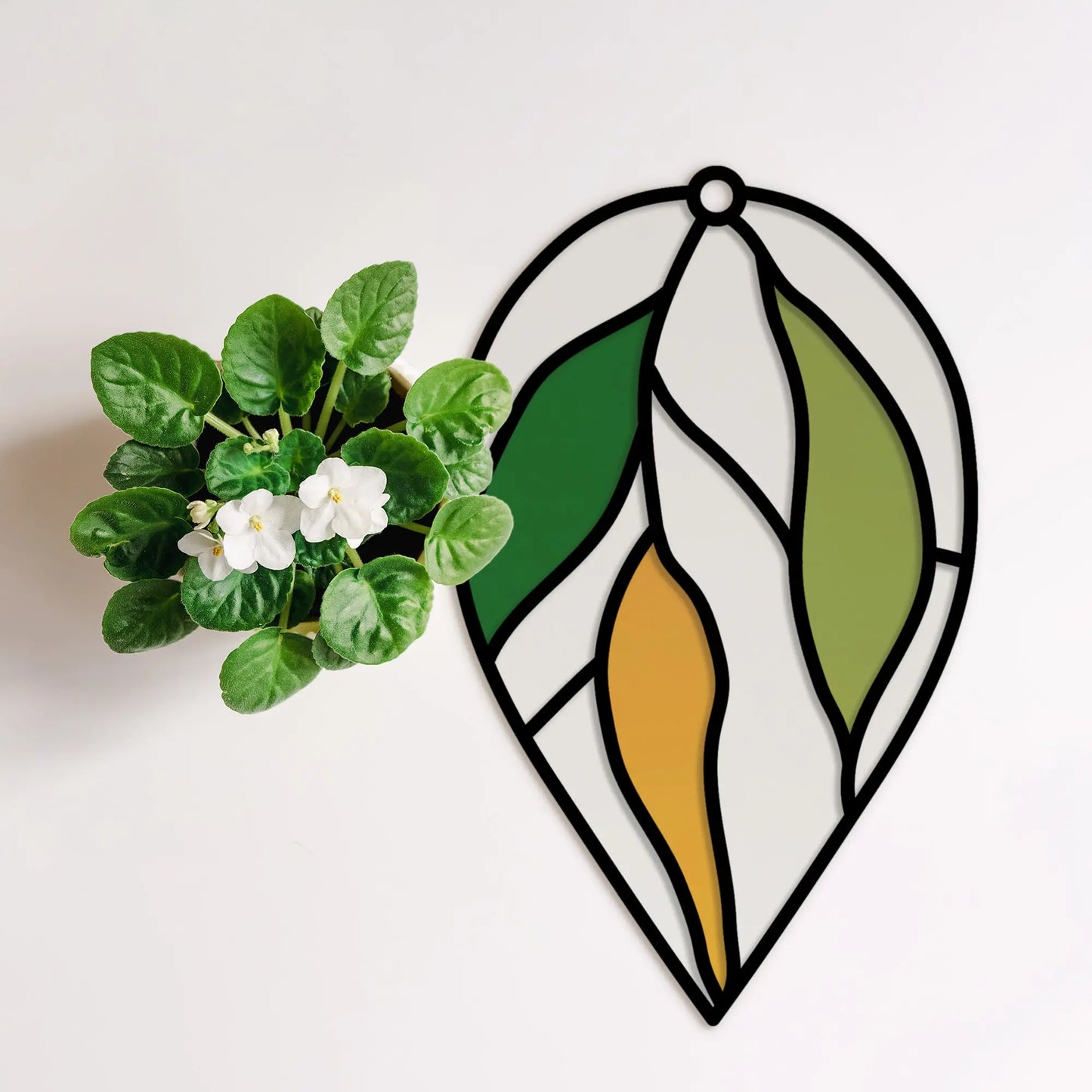 A potted plant with white flowers and green leaves sits gracefully on a white surface beside a stained glass decoration in the shape of a leaf, crafted using the Stained Glass Hanging Leaves Teardrop SVGs from Glowforge, in shades of green, yellow, and white.
