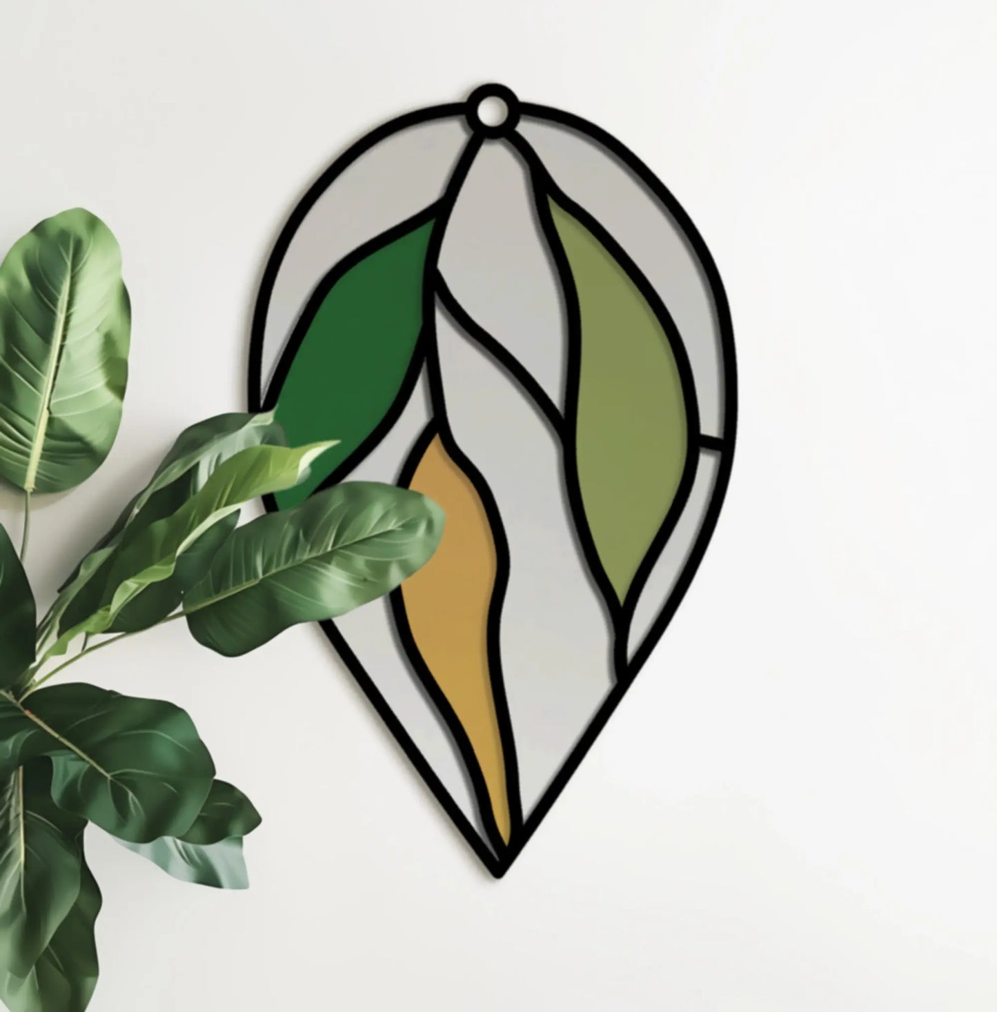 The Stained Glass Hanging Leaves Teardrop SVGs project showcases a decorative glass leaf with green, yellow, and gray sections on a white wall, while a leafy plant peeks in from the left.