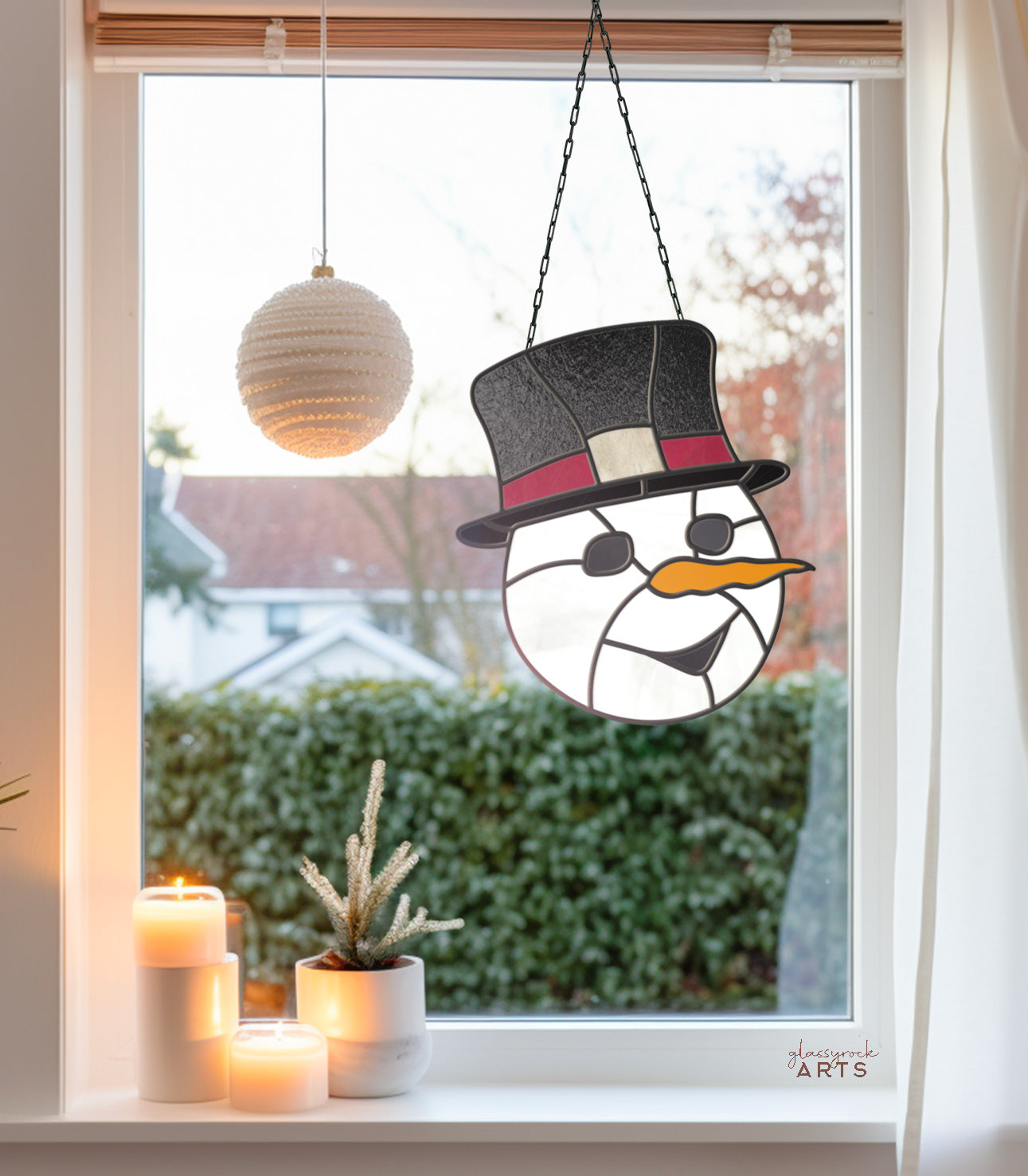 A Snowman Stained Glass Pattern with a black hat and carrot nose hangs in the window, embodying festive Christmas vibes. Nearby, a white spherical ornament dangles. Below, three lit candles and a small potted plant add warmth to the sill, while foliage and houses frame the cozy scene outside.