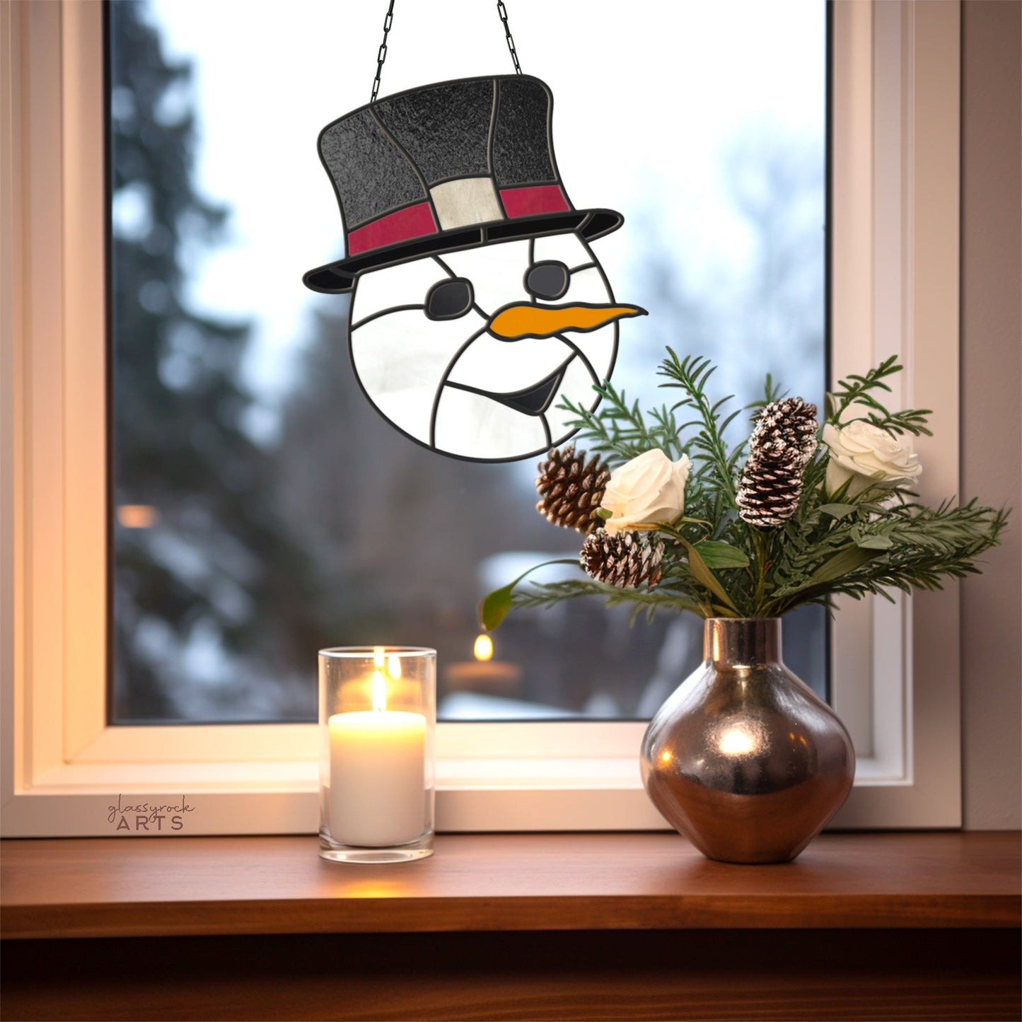 On a window sill, a lit candle sits beside a metallic vase of white flowers and pinecones. Hanging from the window is an exquisite Snowman Stained Glass Pattern ornament. Through the glass, blurry winter trees are faintly visible outside, creating a festive Christmas scene.