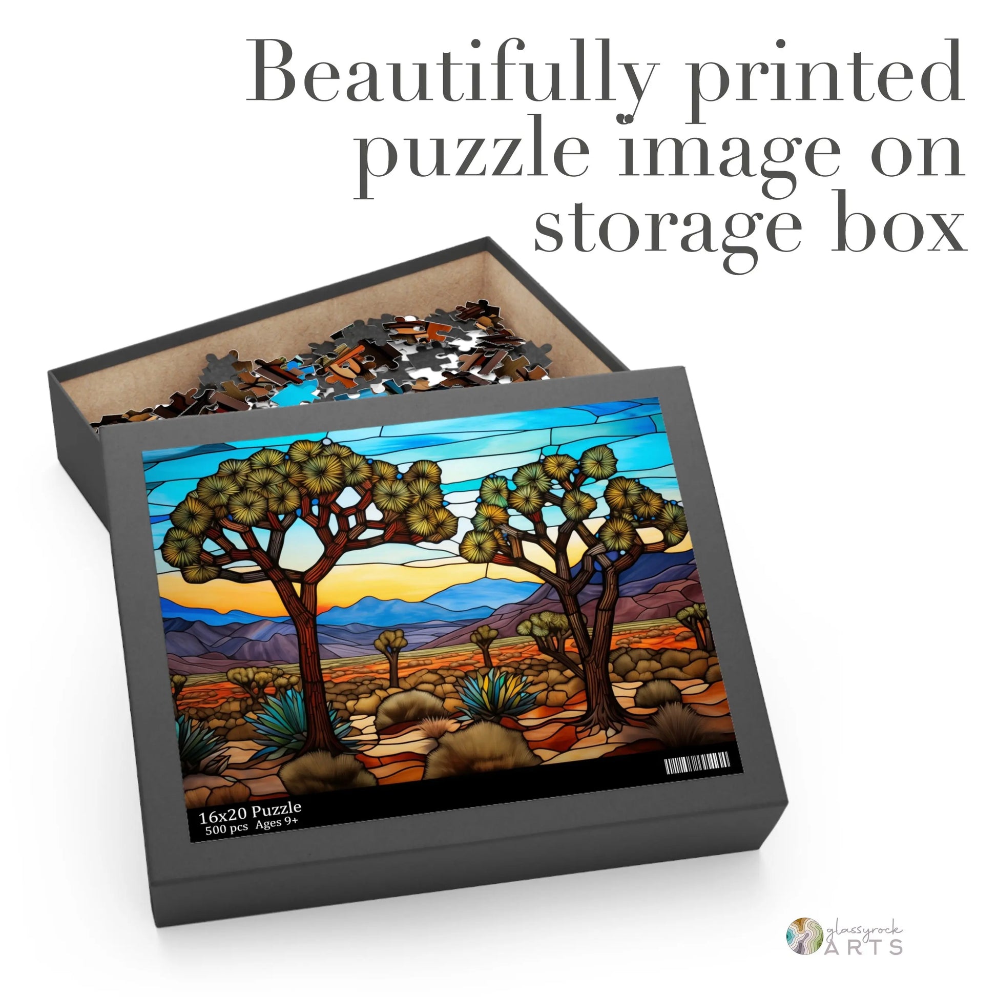 The open box of the Joshua Tree National Park Jigsaw Puzzle reveals pieces showcasing a vibrant stained glass scene of a desert landscape with trees and hills. The boxs text reads: Beautifully printed puzzle image on storage box. Perfect as an art gift.
