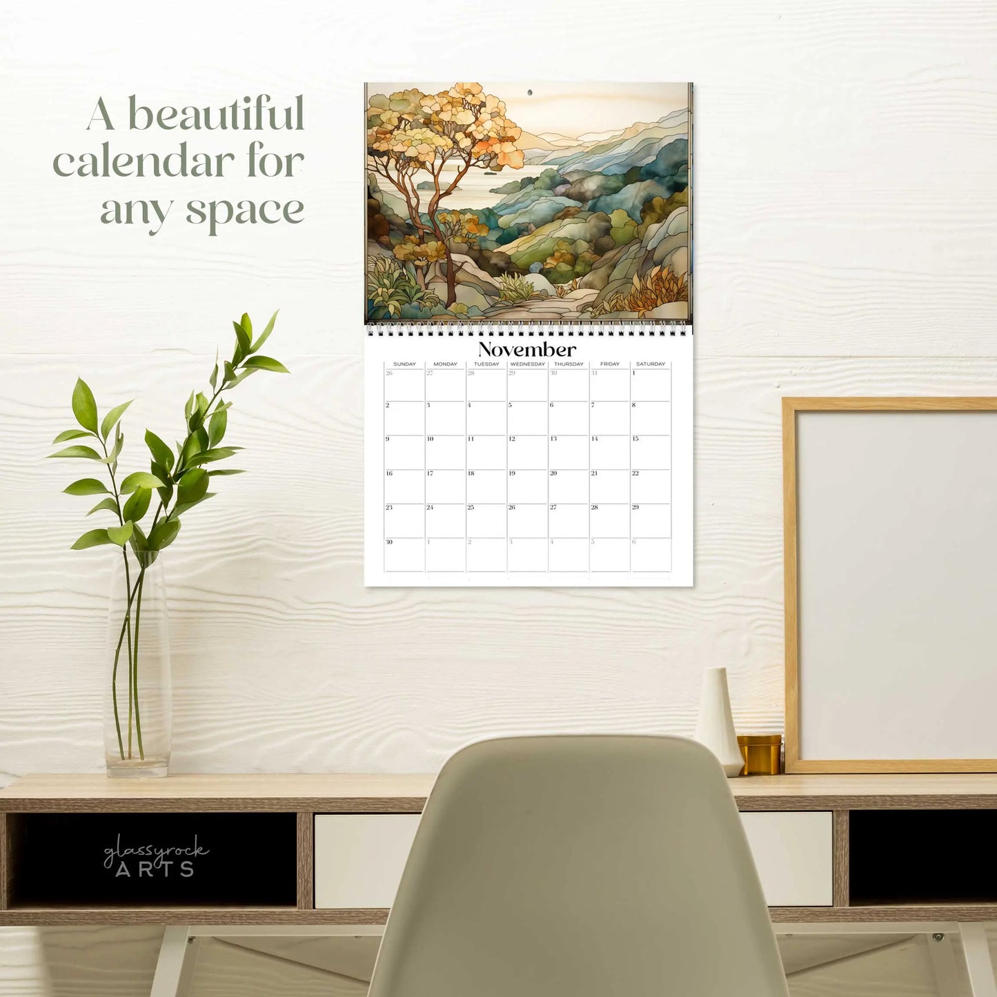 A 2025 Landscapes Stained Glass Wall Calendar open to November hangs above a modern desk, showcasing an artistic nature landscape of trees, hills, and a cloudy sky. The desk features a small plant and framed objects. Text reads, A beautiful calendar for any space.