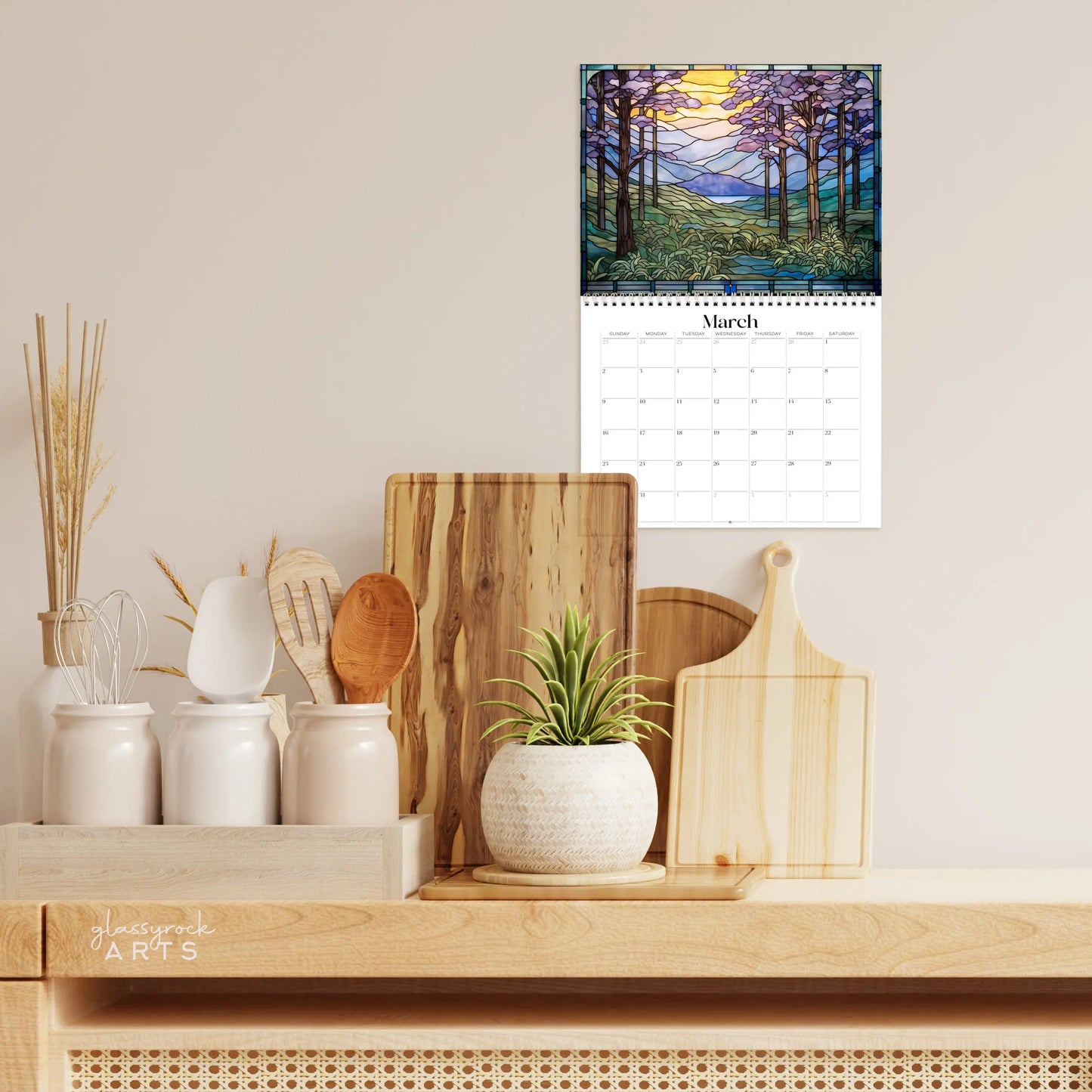 A kitchen counter with decorative cutting boards, a potted succulent, and wooden utensils sits beneath the 2025 Landscapes Stained Glass Wall Calendar, showcasing a nature scene for March, framed by a neutral-toned wall.