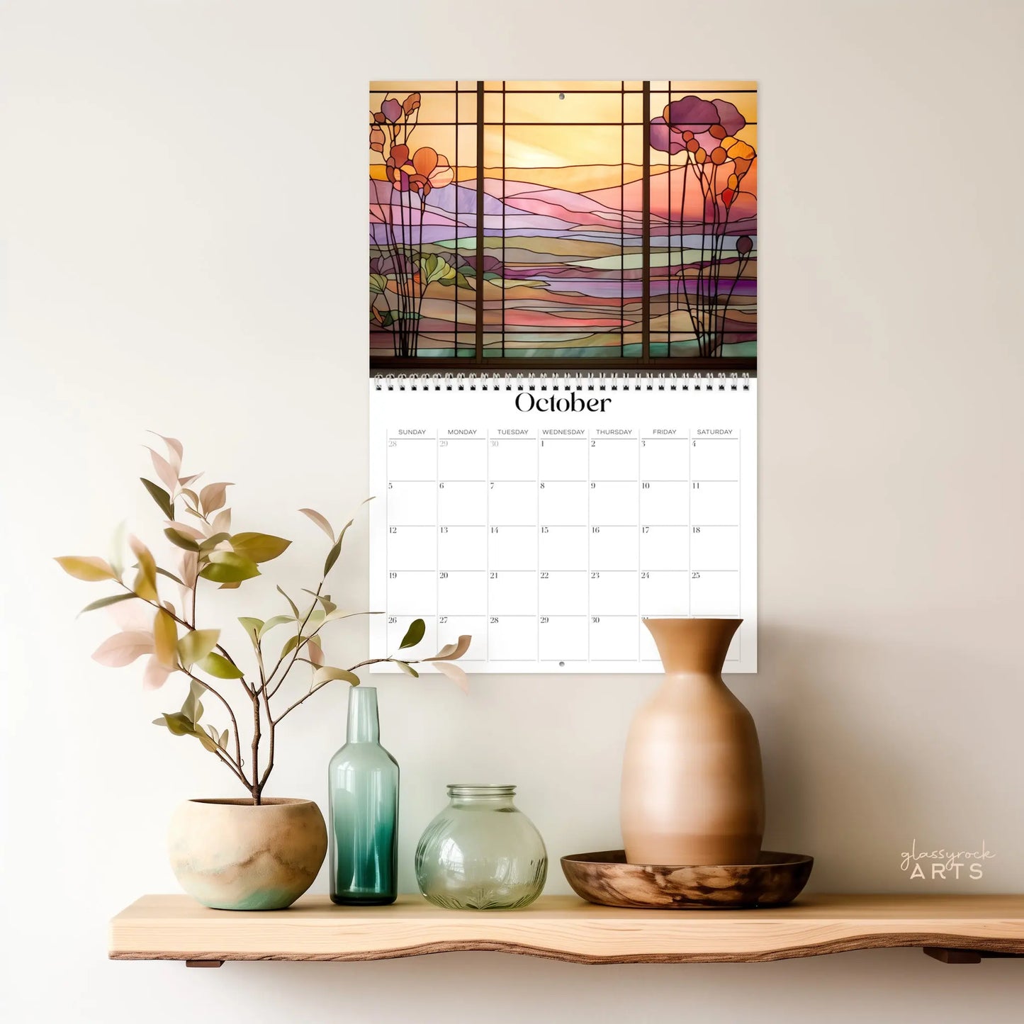 The 2025 Landscapes Stained Glass Wall Calendar, showcasing floral designs like a nature landscape for October, is hung above a wooden shelf adorned with decorative vases and a small plant.