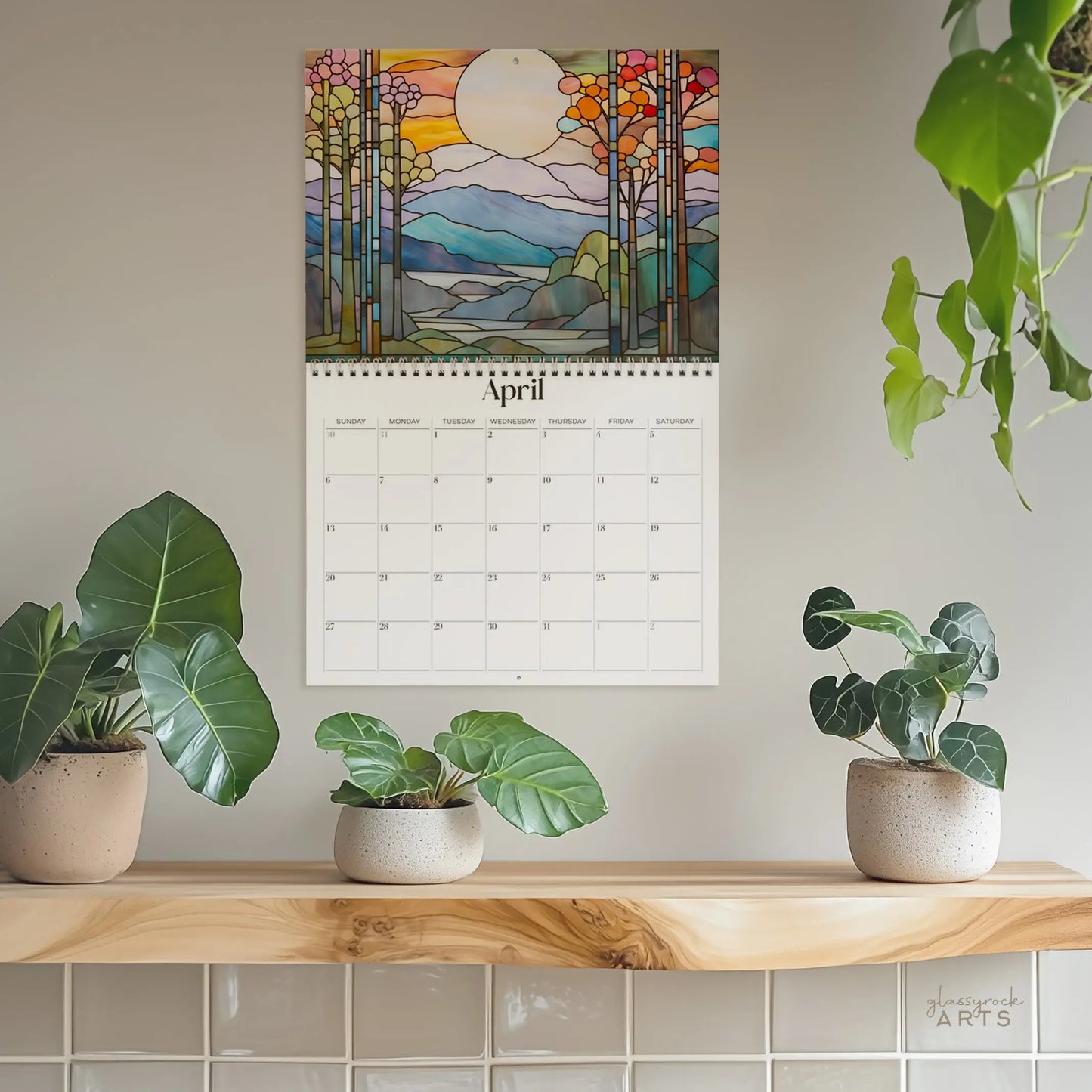 A 2025 Landscapes Stained Glass Wall Calendar opened to April adorns a light wall, displaying a stunning nature scene. Above, art of lush trees and a vivid sun reflects the scene like stained glass. Below, potted plants in round containers sit on a wooden shelf, enhancing the greenery.
