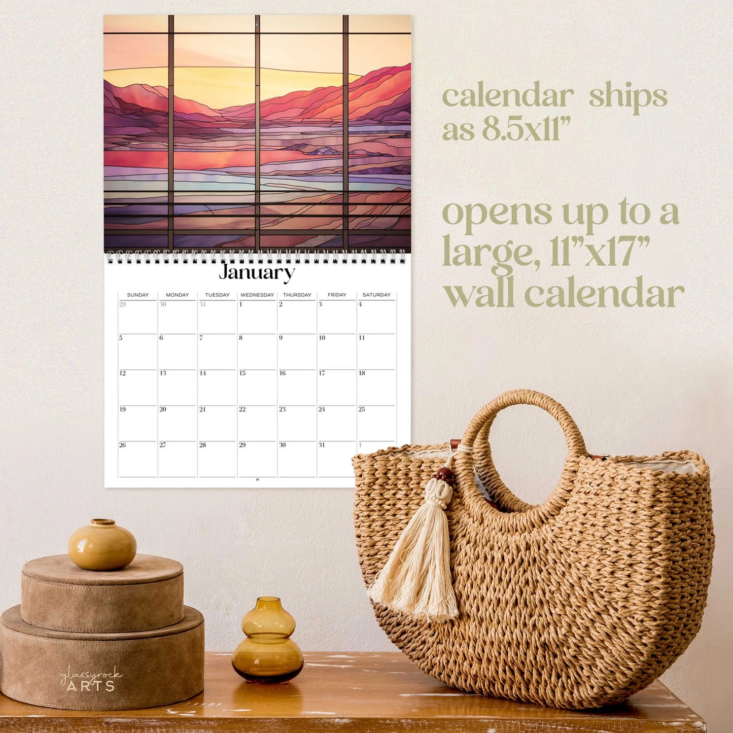The 2025 Landscapes Stained Glass Wall Calendar presents a mountain and lake scene in warm hues, reminiscent of stained glass art. Its elegantly displayed above stacked circular boxes and a woven tassel bag. Text notes the calendar size as 8.5x11 for shipping and 11x17 when opened.