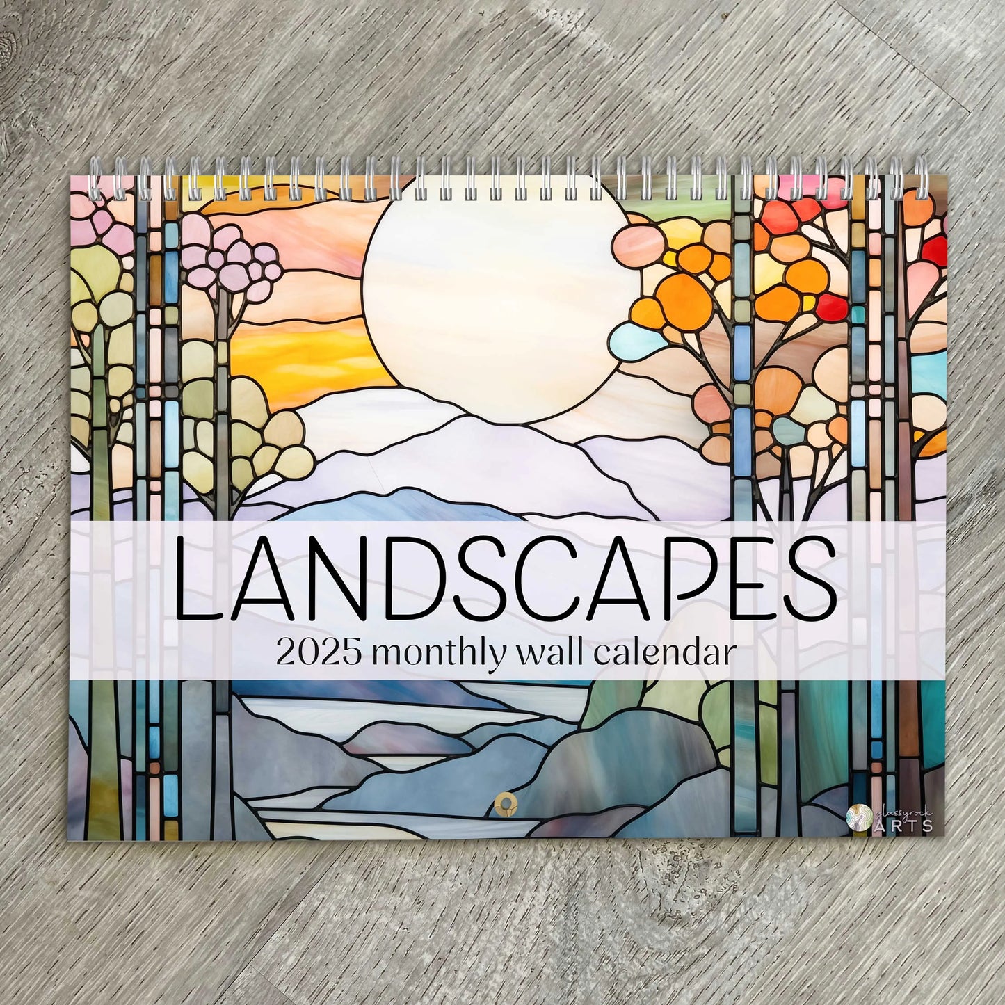 The 2025 Landscapes Stained Glass Wall Calendar displays a vibrant sun, colorful trees, and mountains on its cover. It is spiral-bound and beautifully rests on a wooden surface.