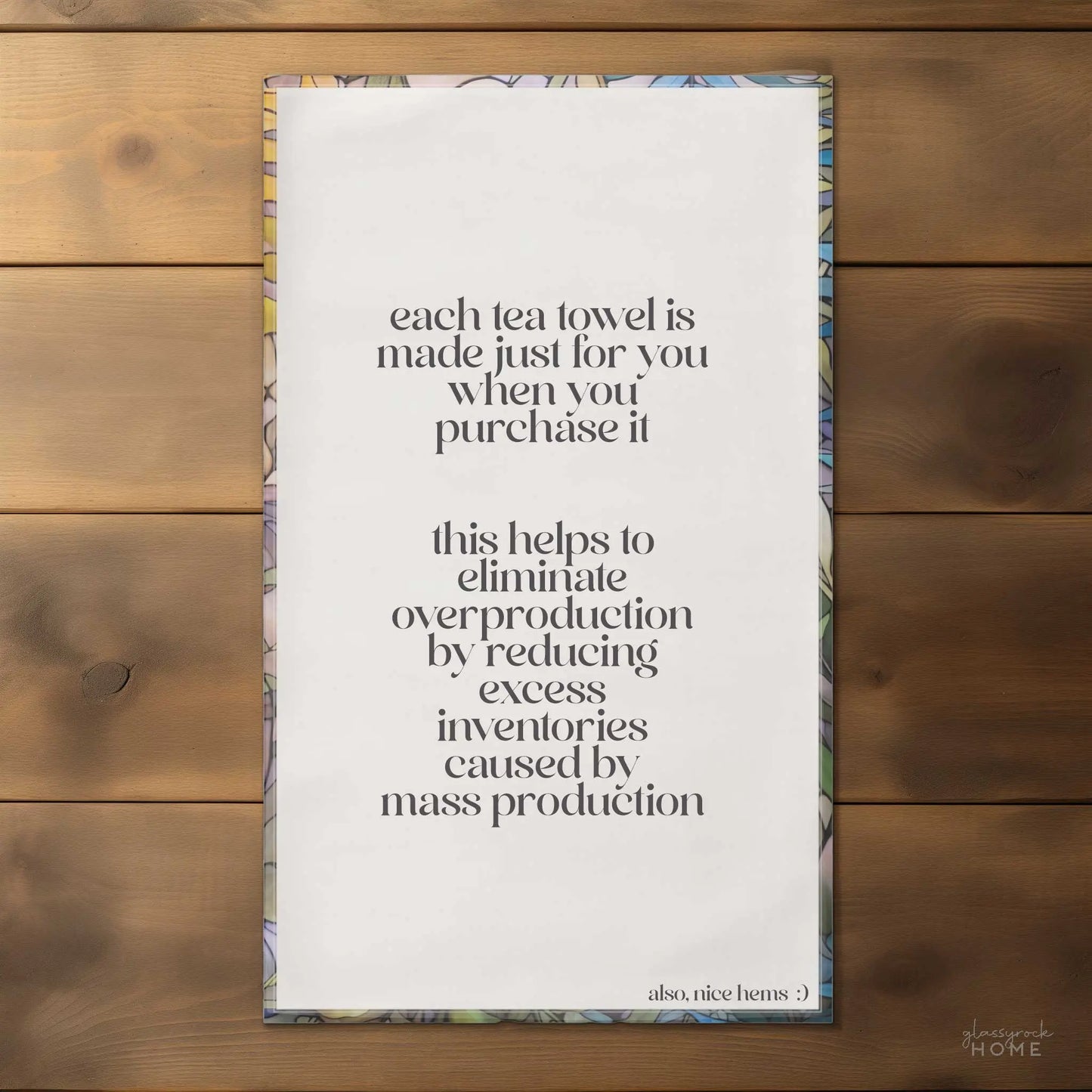 The Stained Glass Pastel Ferns Dish Towel, crafted by a Pacific Northwest artist, highlights sustainability with the message: Each towel is made just for you when purchased, reducing overproduction and excess inventory. Also, nice hems :) on a wooden surface.