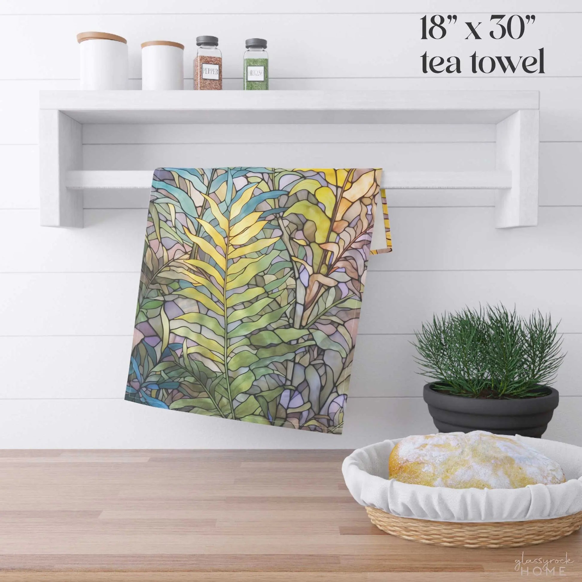 The Stained Glass Pastel Ferns Dish Towel, featuring a leafy design, hangs on a white shelf. It sits beneath two white canisters and adorable jars. A yellow-white cloth basket beside a potted plant completes this charming setup. Text reads 18 x 30 tea towel.