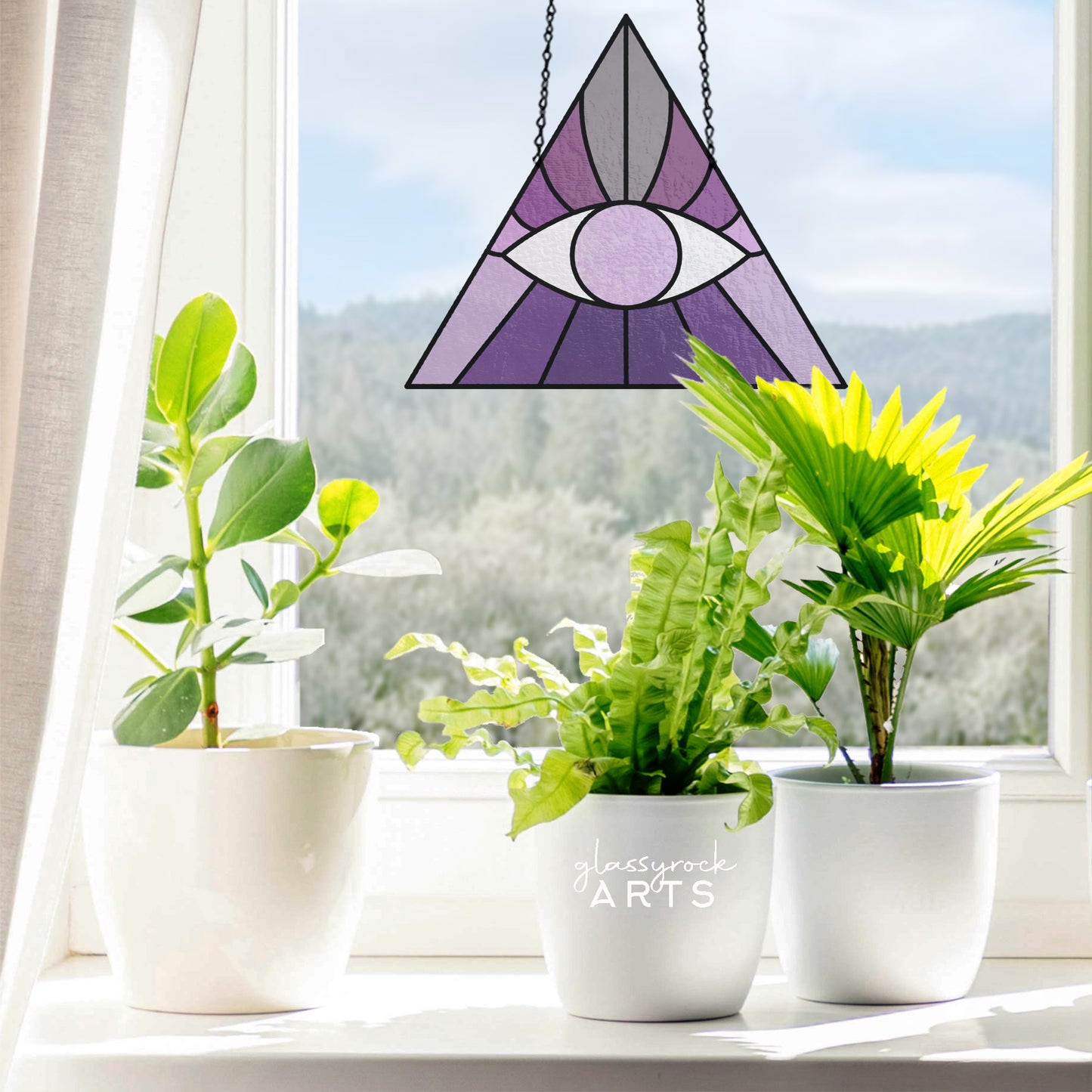 Three potted plants line the windowsill with a scenic view of trees and hills. The Boho Eye Triangle Stained Glass Pattern, featuring a purple eye design, hangs in the window. Sunlight streams through, illuminating both the plants and the vibrant glass art.
