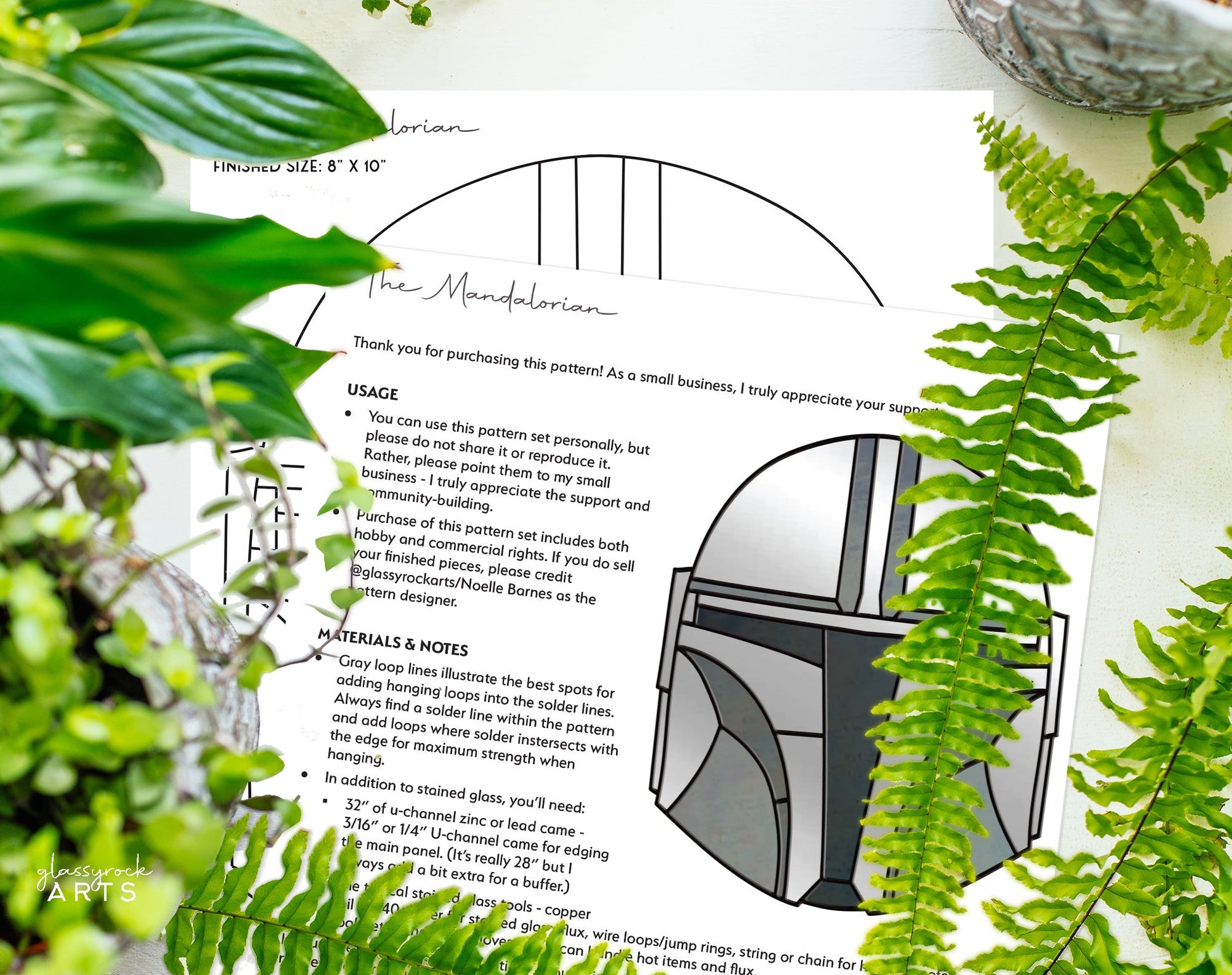 Amid vibrant green ferns, a printed pattern titled The Mandalorian Star Wars Stained Glass Pattern is shown with its instructions and materials. A partial image of the digital design appears on the paper, accompanied by a small gray figurine.